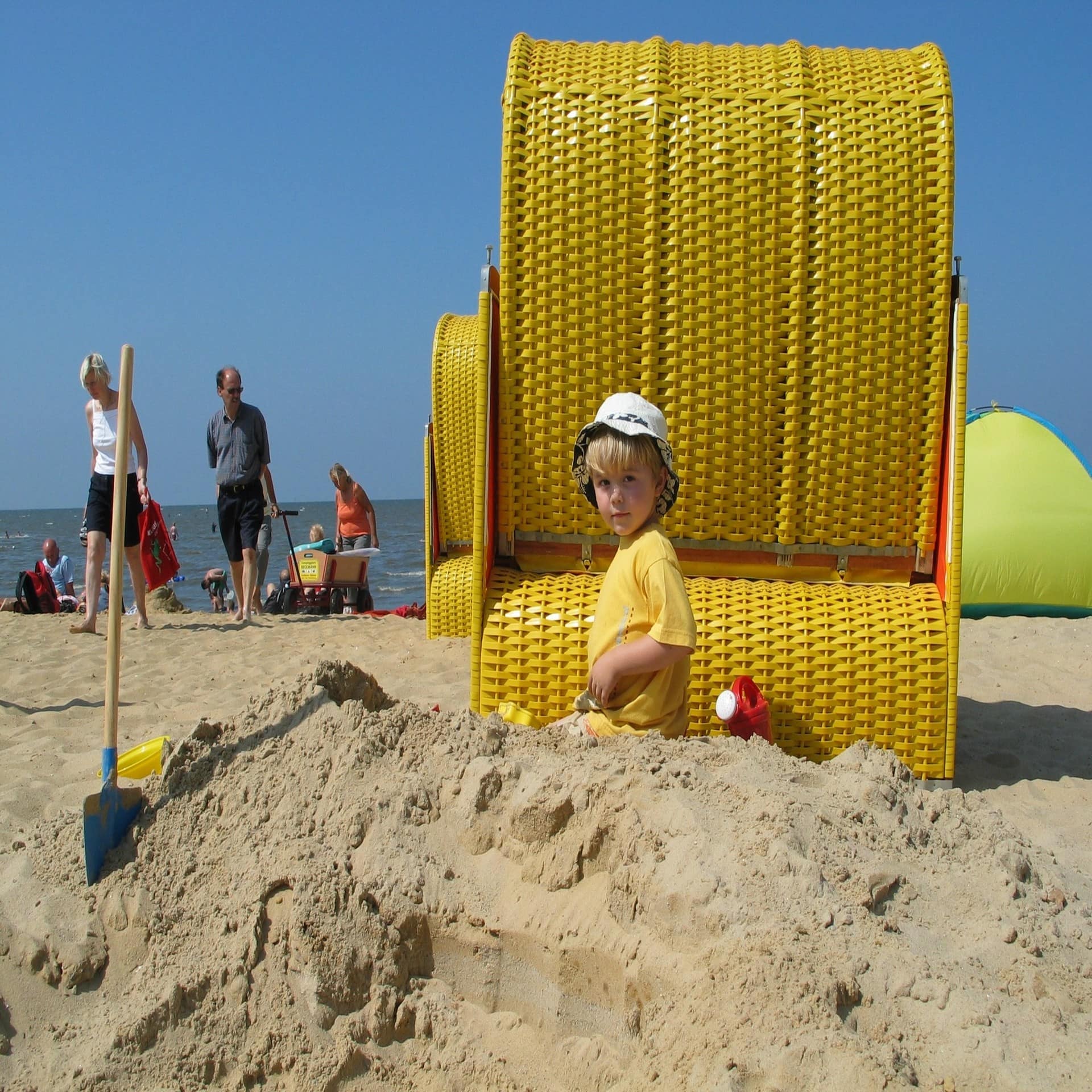 Ein kleiner Junge mit gelbem T-Shirt und Sonnenhut spielt hinter einem gelben Strandkorb im Sand. Links daneben sind Leute. 