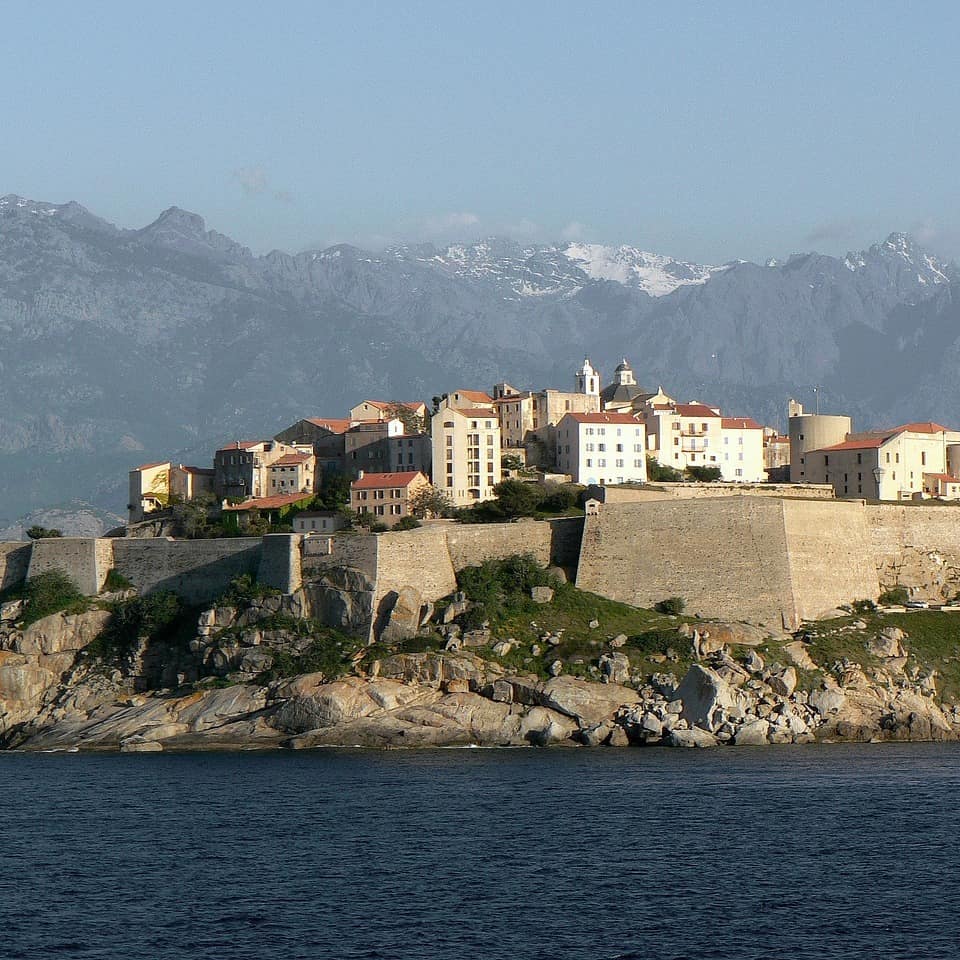 Blick vom Wasser auf Calvi, eine auf einem Hügel liegende Stadt, die von einer Mauer umgeben ist.