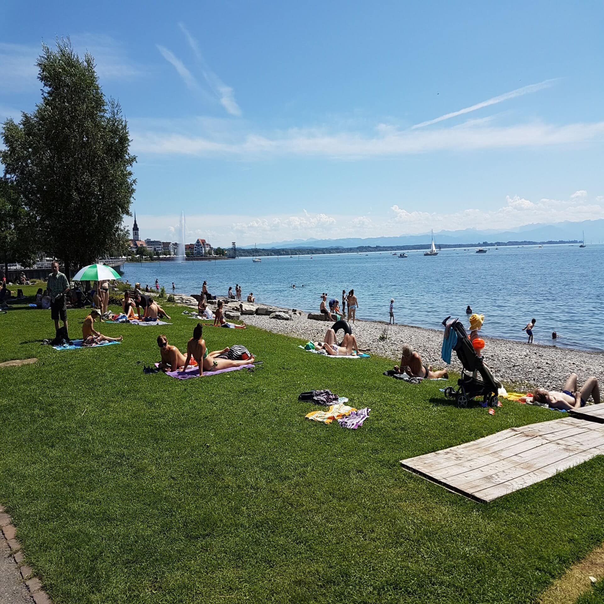 Leute sonnen sich auf einer Liegewiese mit Strand am Bodensee, links ist ein Ort zu sehen.