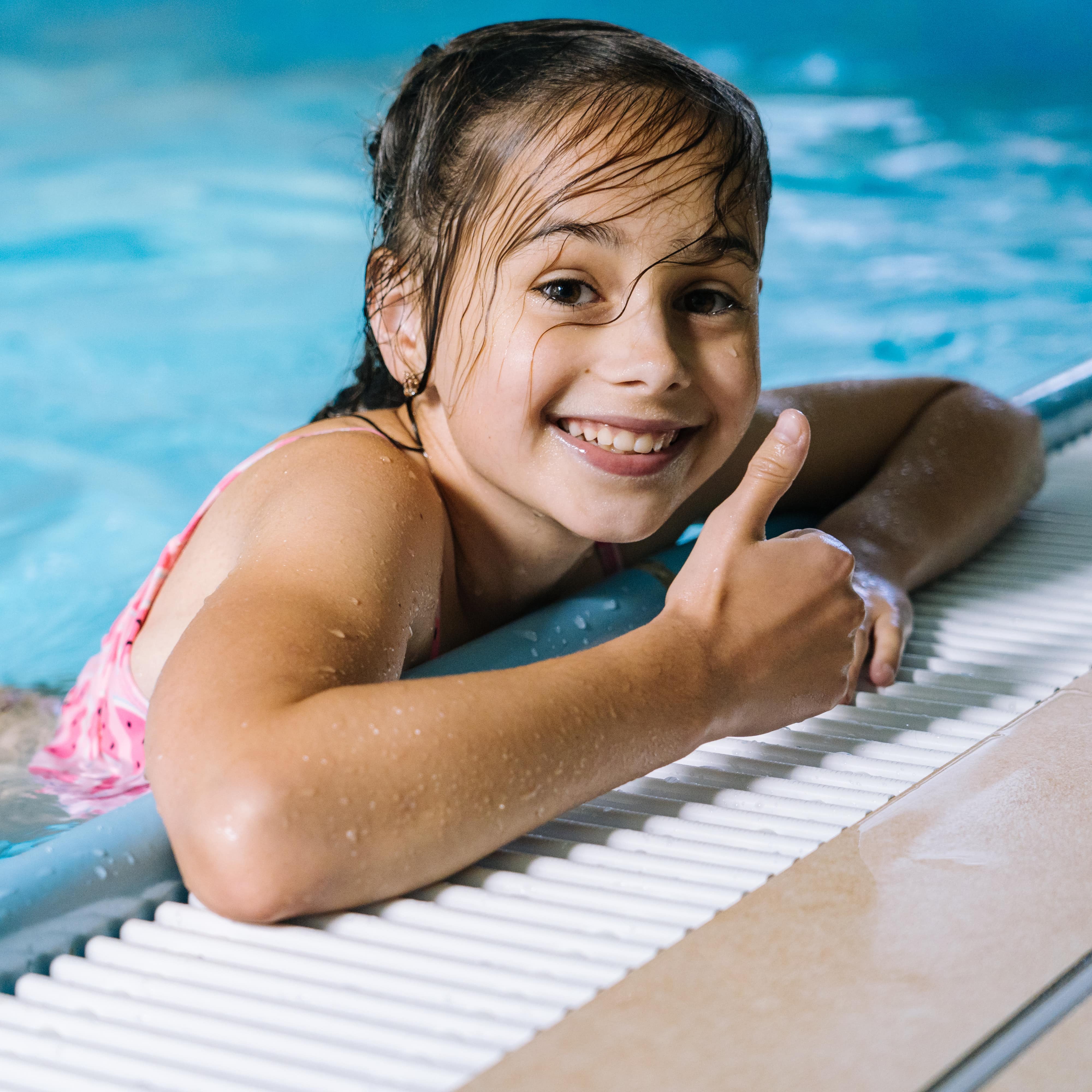 Mädchen im Wasser am Beckenrand eines Pools lacht in die Kamera und streckt den Daumen hoch.