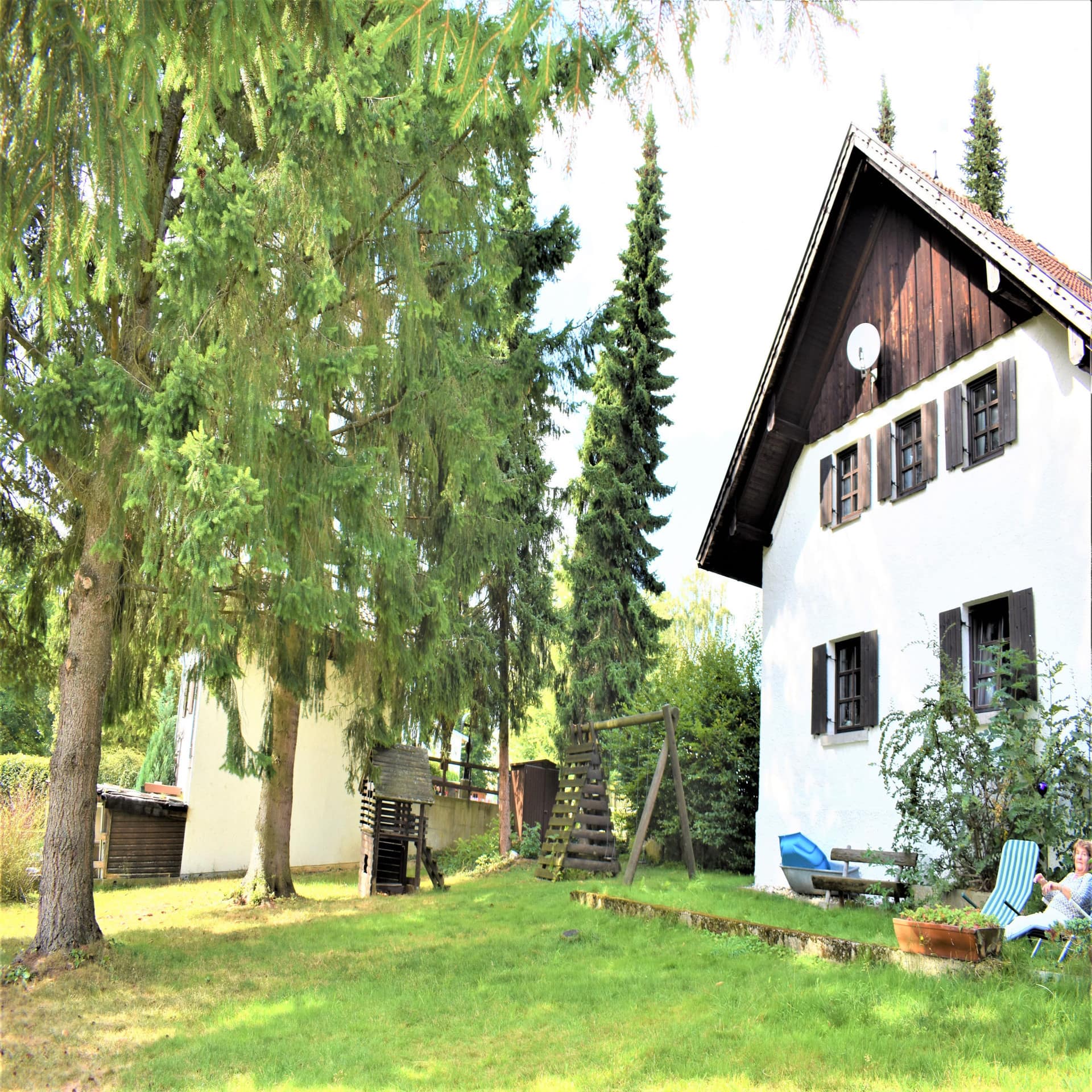 Weißes Bauernhaus mit Garten und kleinem Spielplatz, umgeben von Bäumen.