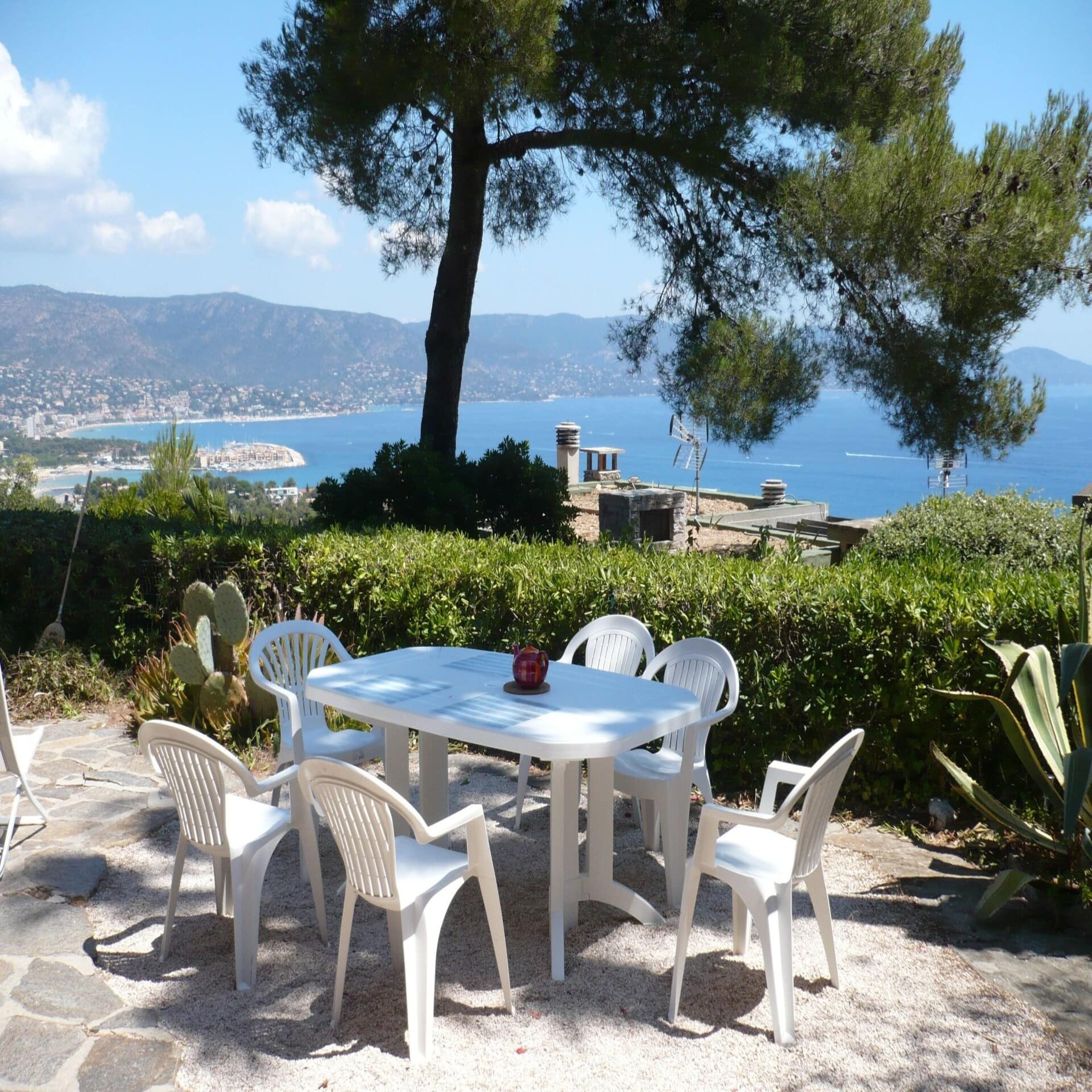 Weißer Tisch mit 6 Stühlen auf einer Terrasse, mit Blick auf den Wasser und einen Ort. 