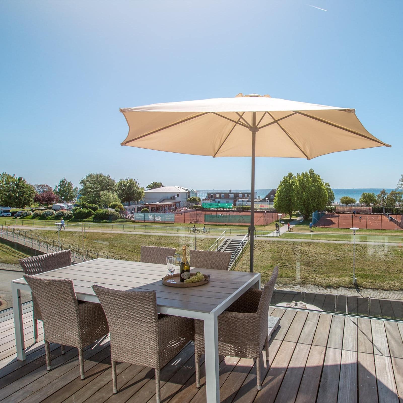 Balkon mit Glas, Tisch, Stühlen und Sonnenschirm sowie Blick auf Tennisplätze und die Ostsee