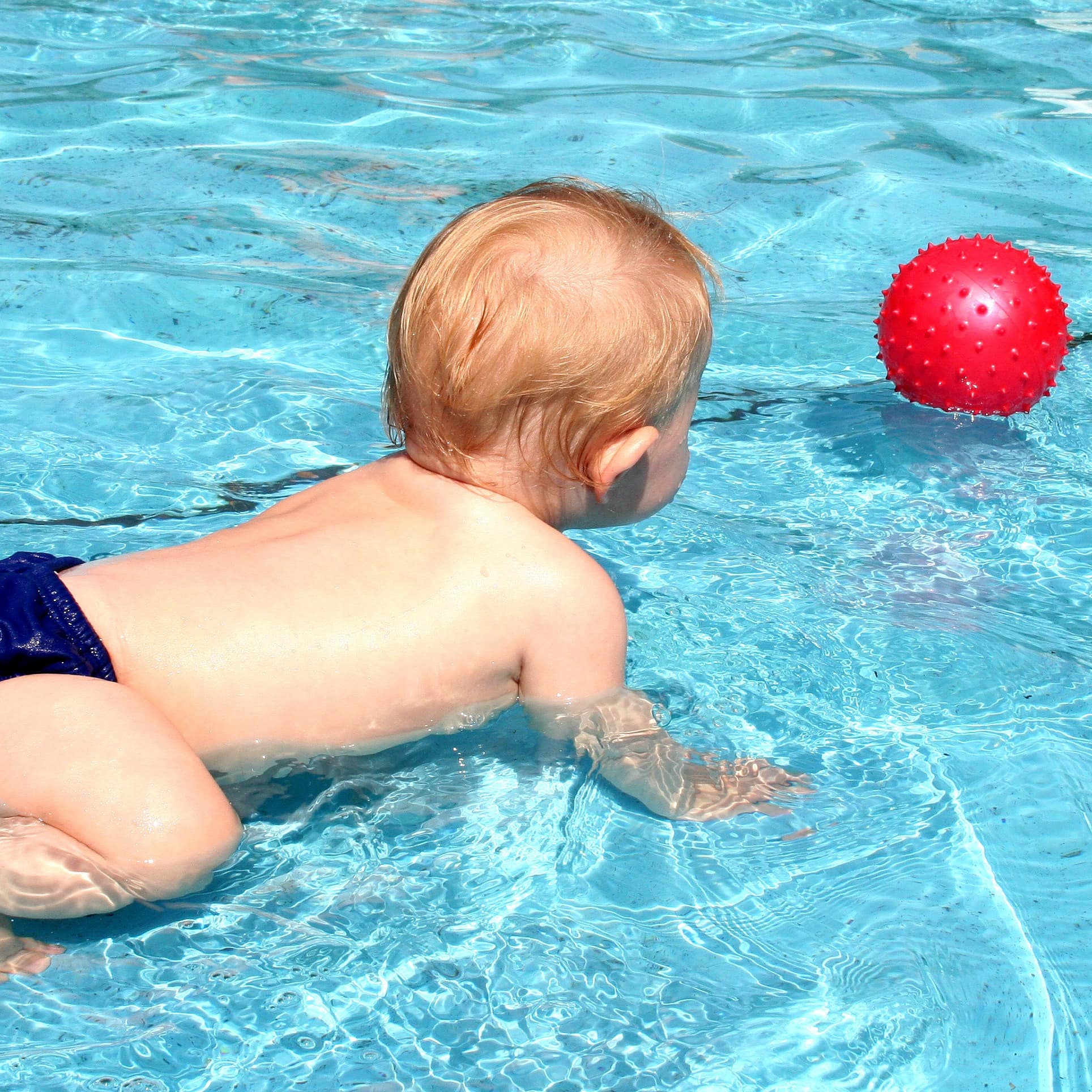 Baby mit blauer Badehose krabbelt im flachen Wasser eines Pools auf einen roten Ball zu.