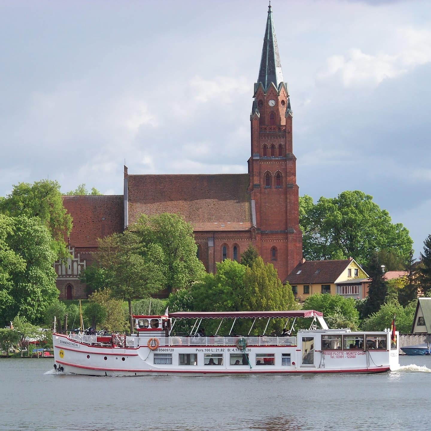 Blick über das Wasser auf eine Kirche und Häuser von Müritz, auf dem Wasser fährt ein Boot vorbei