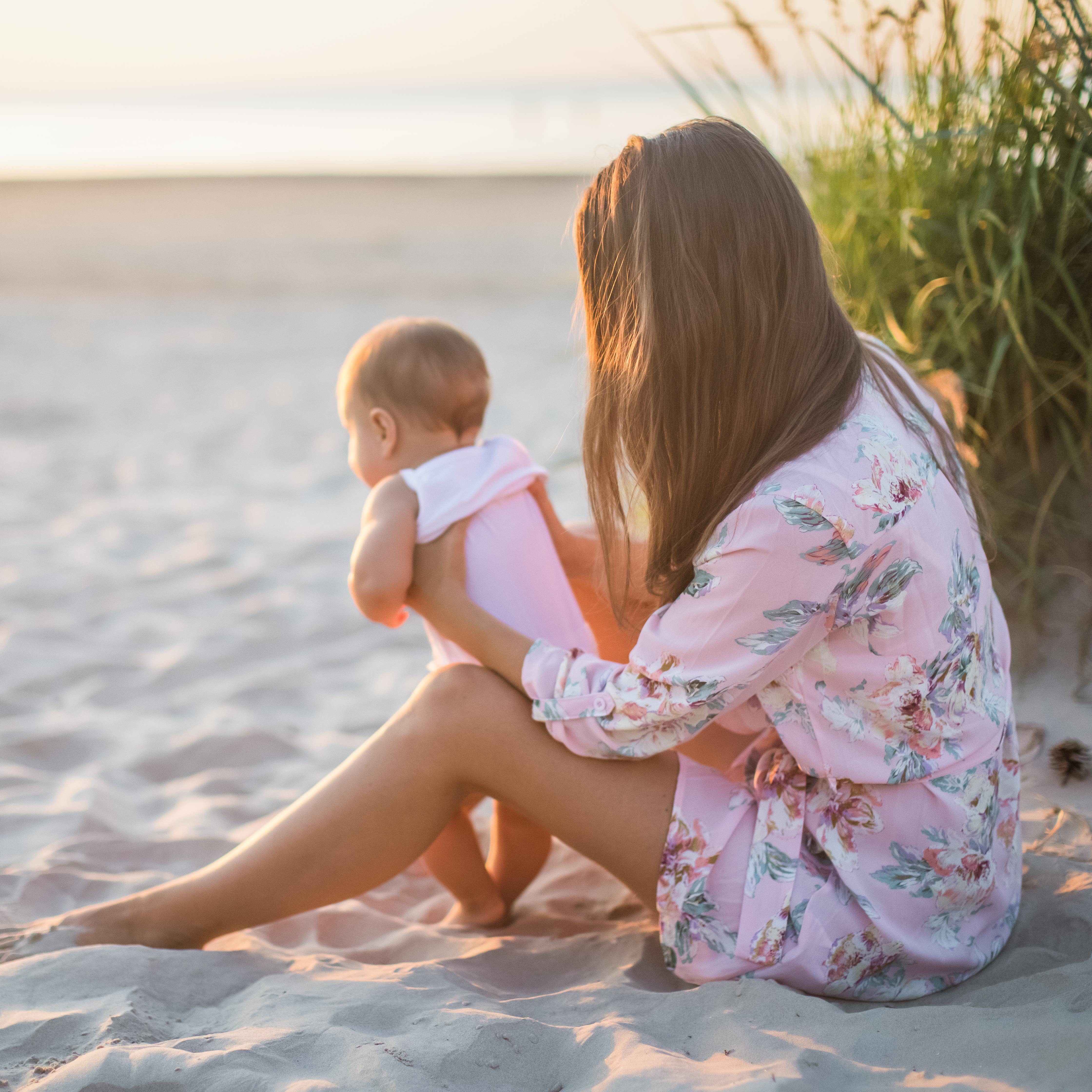 Blick von der Seite: Eine Mutter im rosa Blumenkleid hält ein Baby im Strampler am Strand, daneben Dünengras.