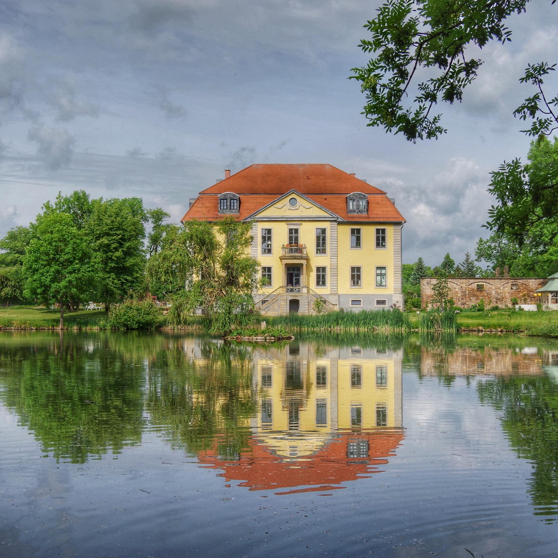 Ein kleines Schloss spiegelt sich im davorliegenden See. Ringsherum Bäume.