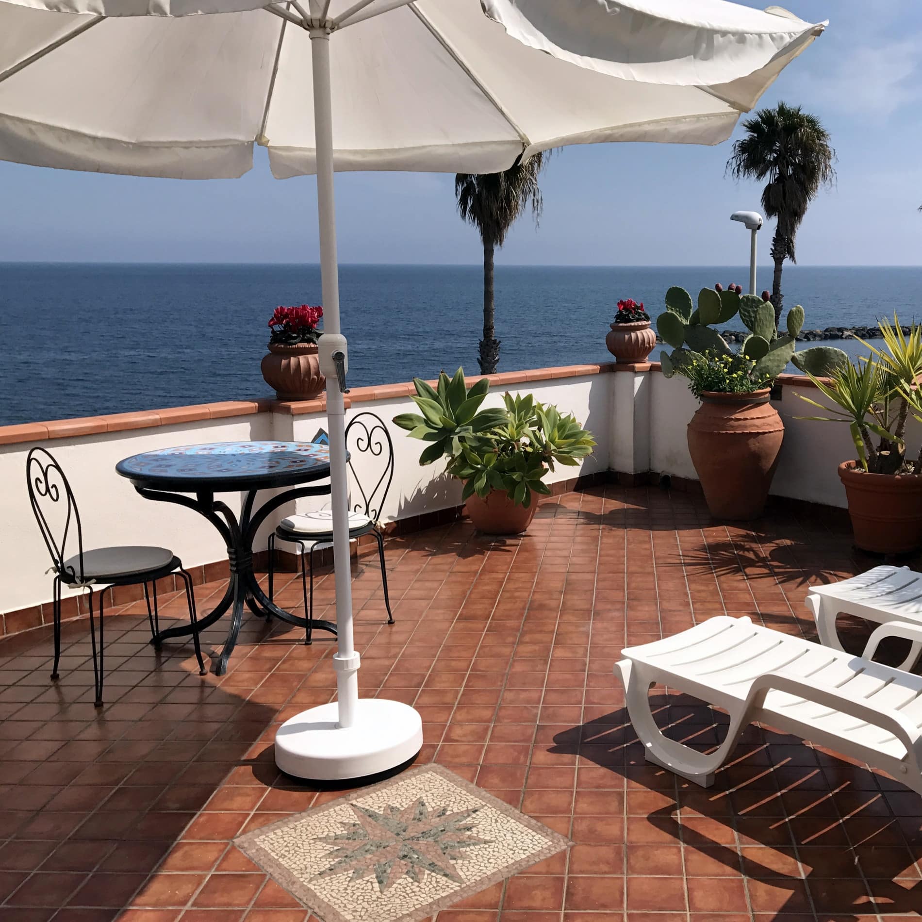 Zwei Sonnenliegen unter einem Sonnenschirm auf der Terrasse der Ferienwohnung in Sizilien blicken hinaus aufs Ionische Meer