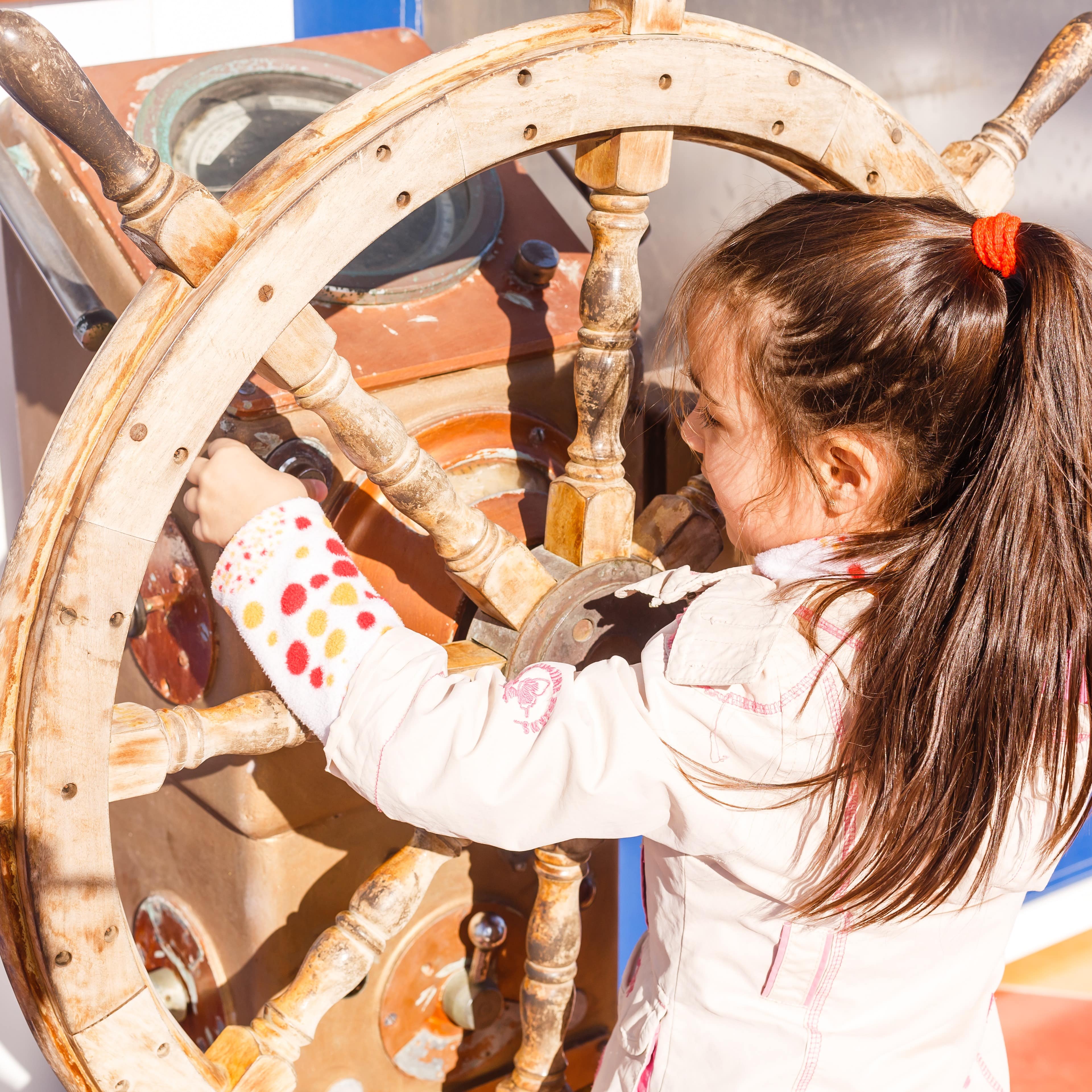 Kleines Mädchen mit langem braunen Zopf dreht am Steuerrad eines Schiffs. 