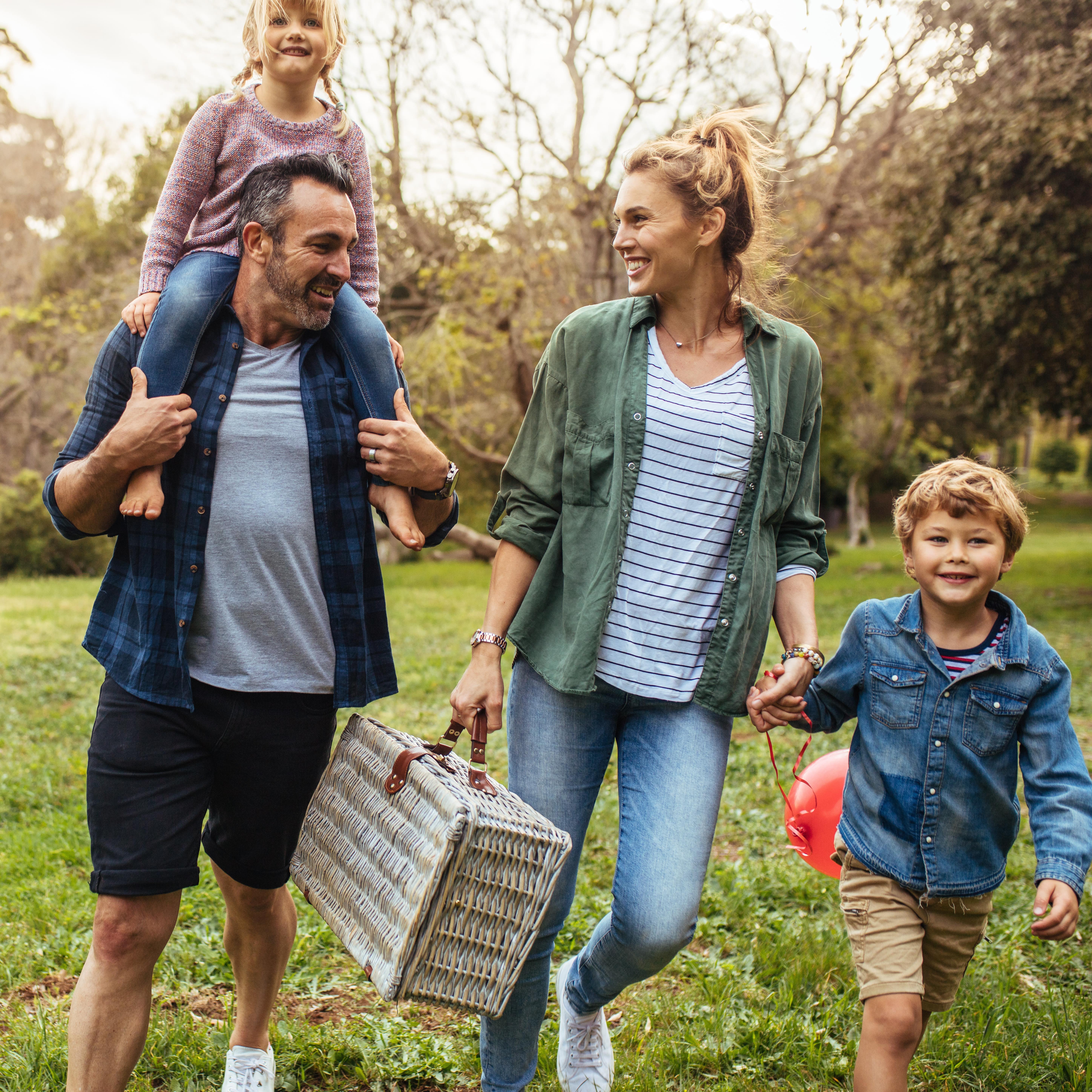 Familie mit 2 Kindern, Hund und Picknickkorb im Grünen auf dem Weg zu Picknick.