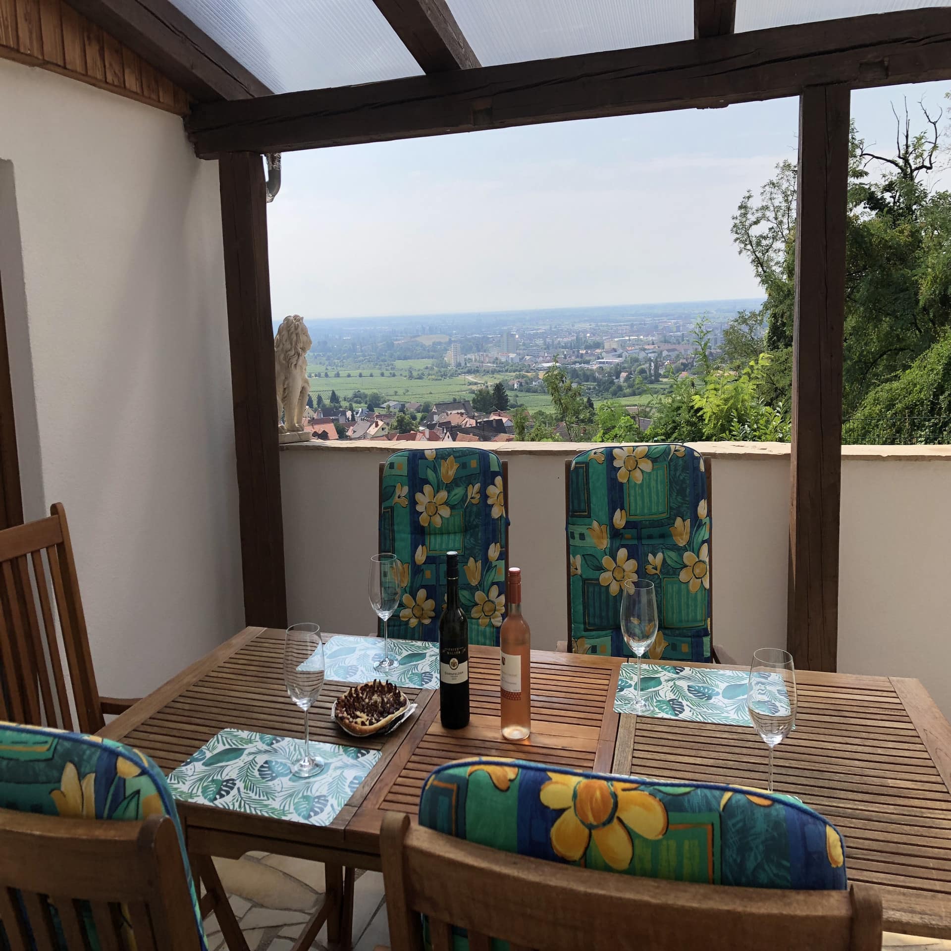 Balkon mit Tisch auf dem 2 Weinflaschen, Weingläser und etwas zu Essen stehen. Aussicht auf die Weinberge und Häuser.
