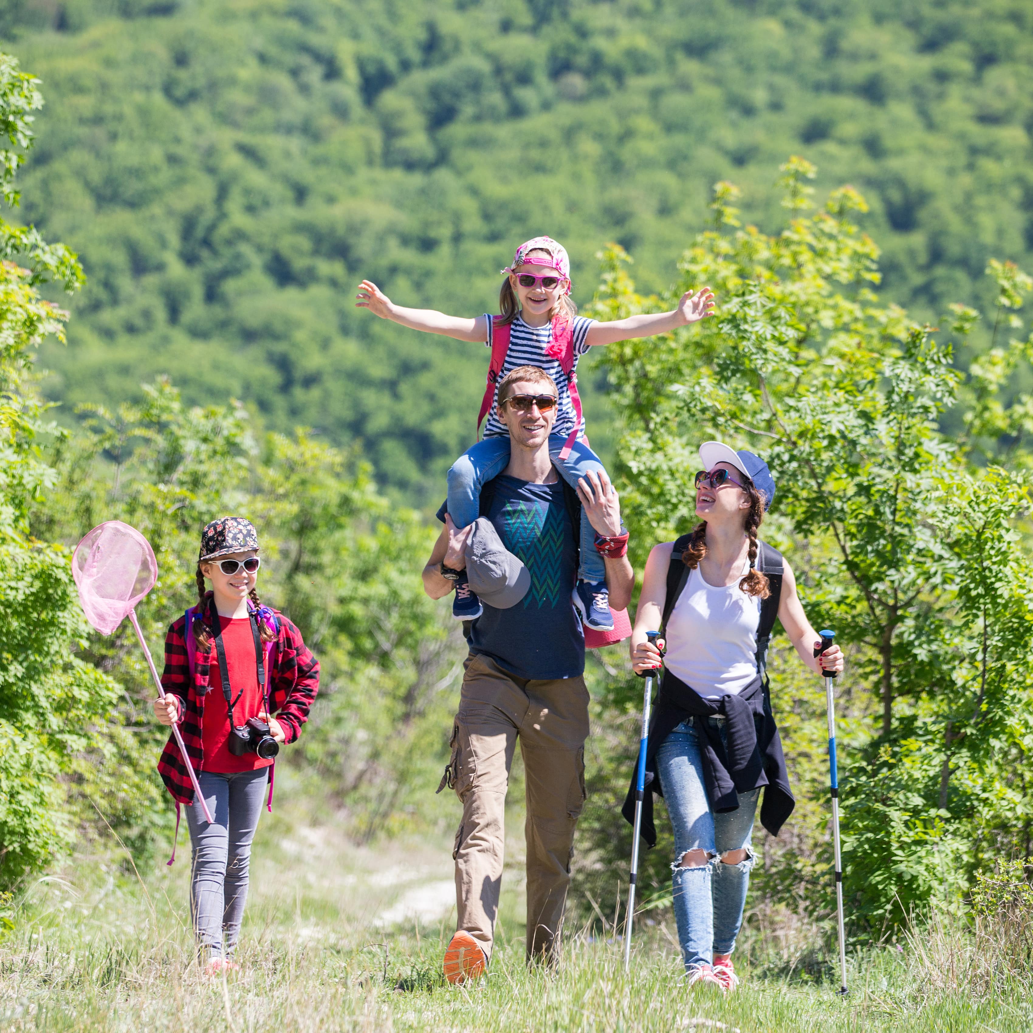 Familie mit 2 Töchtern wandert durch die grüne Natur, alle tragen Sonnenbrillen.