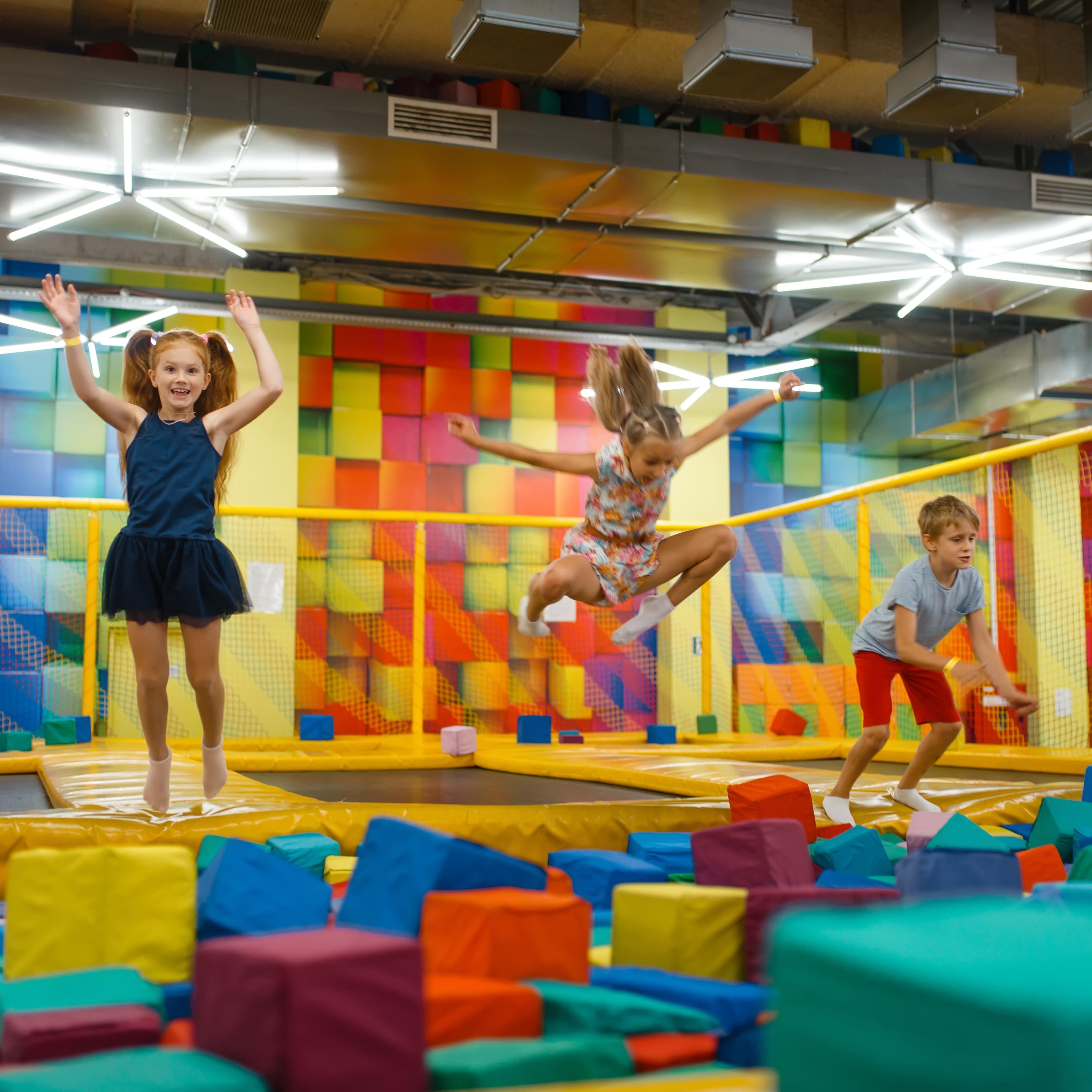 3 Kinder springen auf Trampolinen in einem bunten Trampolinpark.