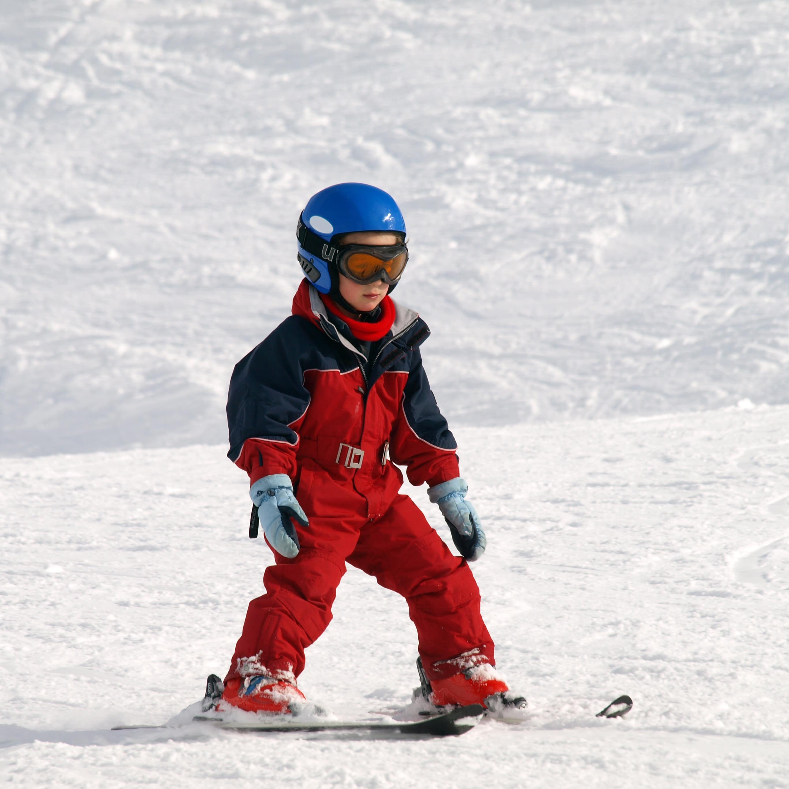 Kleiner Junge in rotem Schneeanzug und mit blauem Helm fährt Ski. 