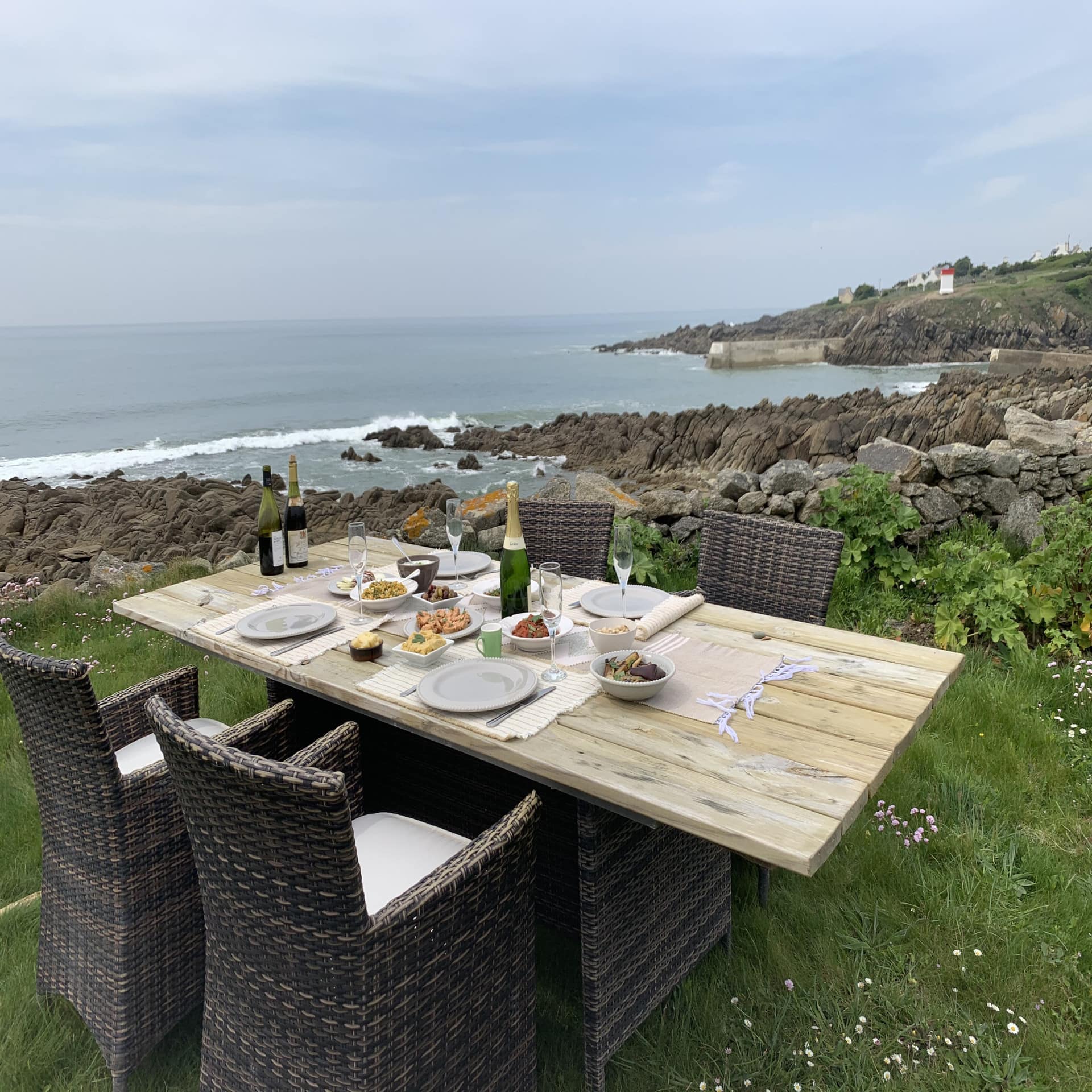 Gedeckter Tisch für 4 Personen mit Essen Champagner in einem grünen Garten direkt an der felsigen Küste an Meer.