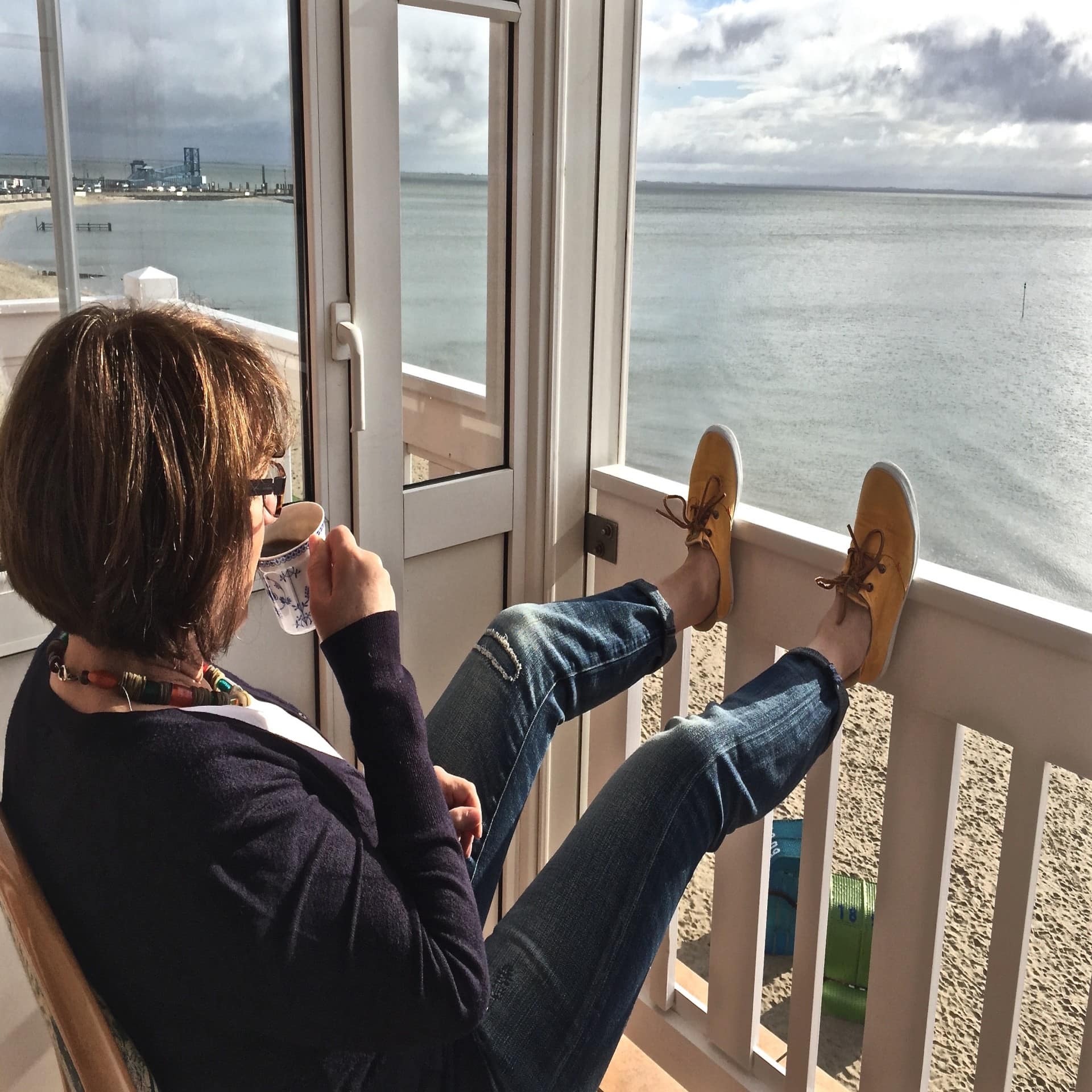 Frau mit Kaffeetasse sitzt auf einem Balkon direkt am Strand und blickt aufs Meer, unten stehen 2 Strandkörbe. 