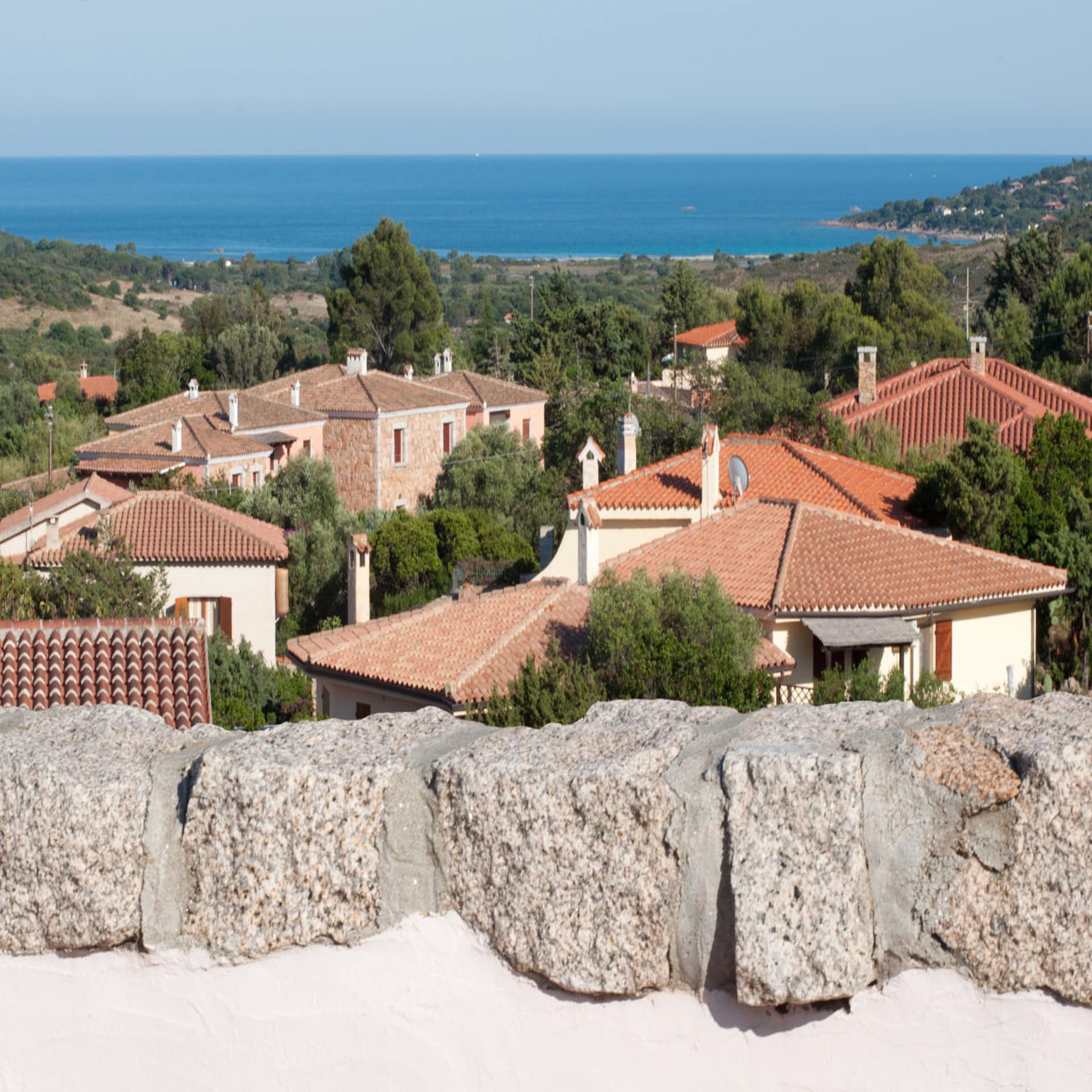Blick von einem Balkon über weiße Häuser mit roten Dächern auf Sardinien, im Hintergrund das Meer. 