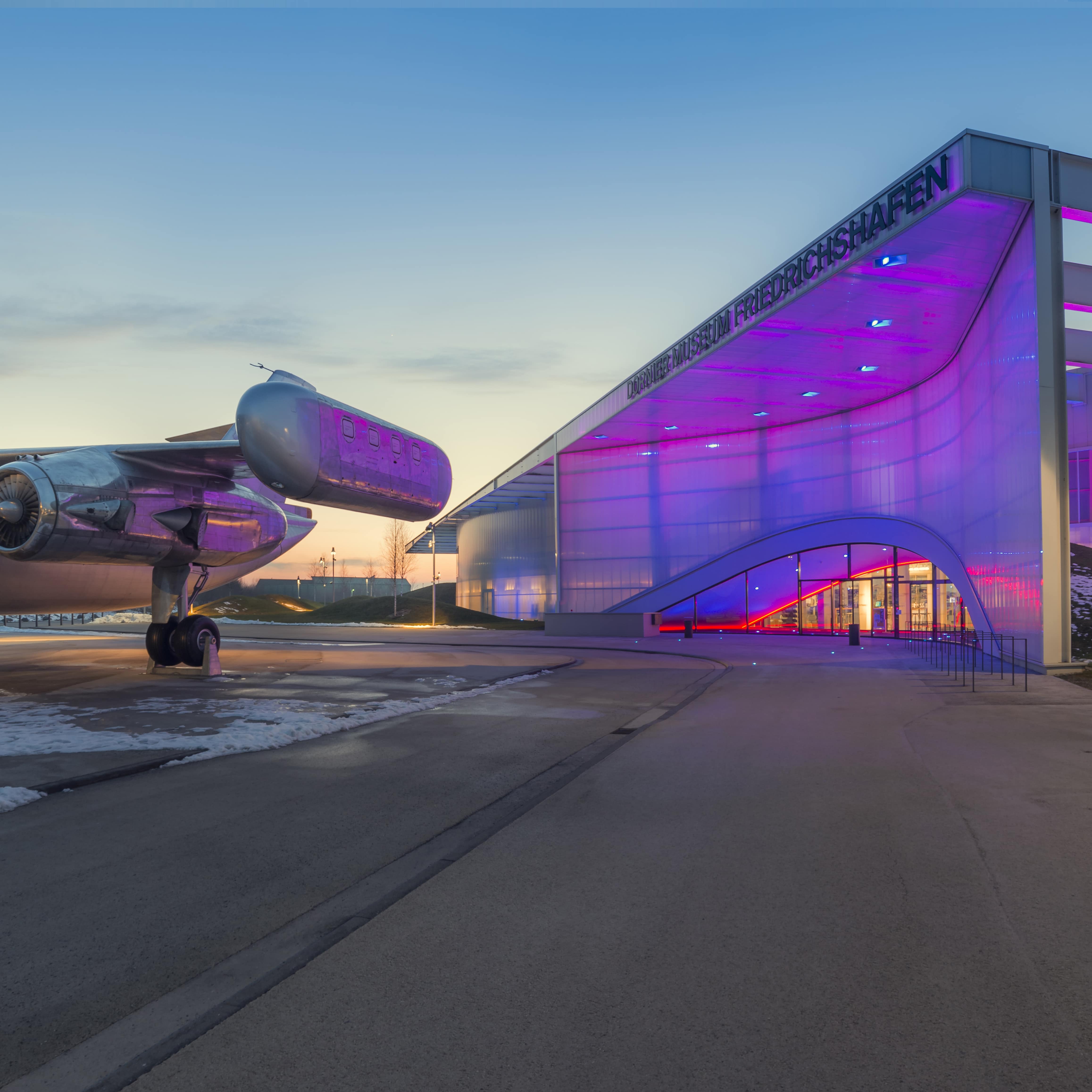 Blick aufs Dornier Museum bei Sonnenuntergang im Winter. Links steht ein Flugzeug, das Gebäude daneben ist lila erleuchtet.