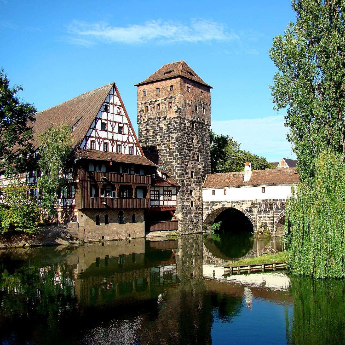 Blick über einen Fluss in Nürnberg, zu sehen sind ein Fachwerkhaus, ein historischer Turm mit Brücke und Bäume