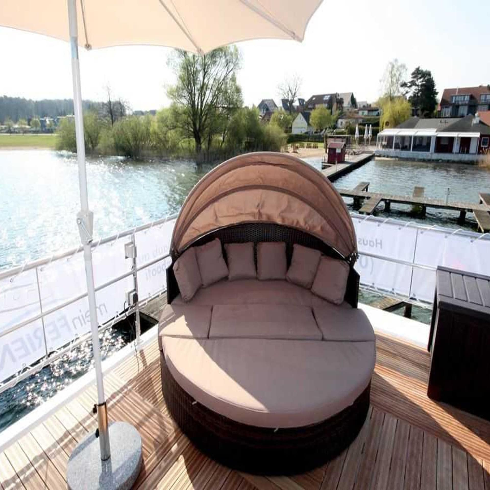 Rundes Sonnenbett mit Sonnenschirm au einem Hausboot das an einem Bootssteg liegt; Häuser im Hintergrund.