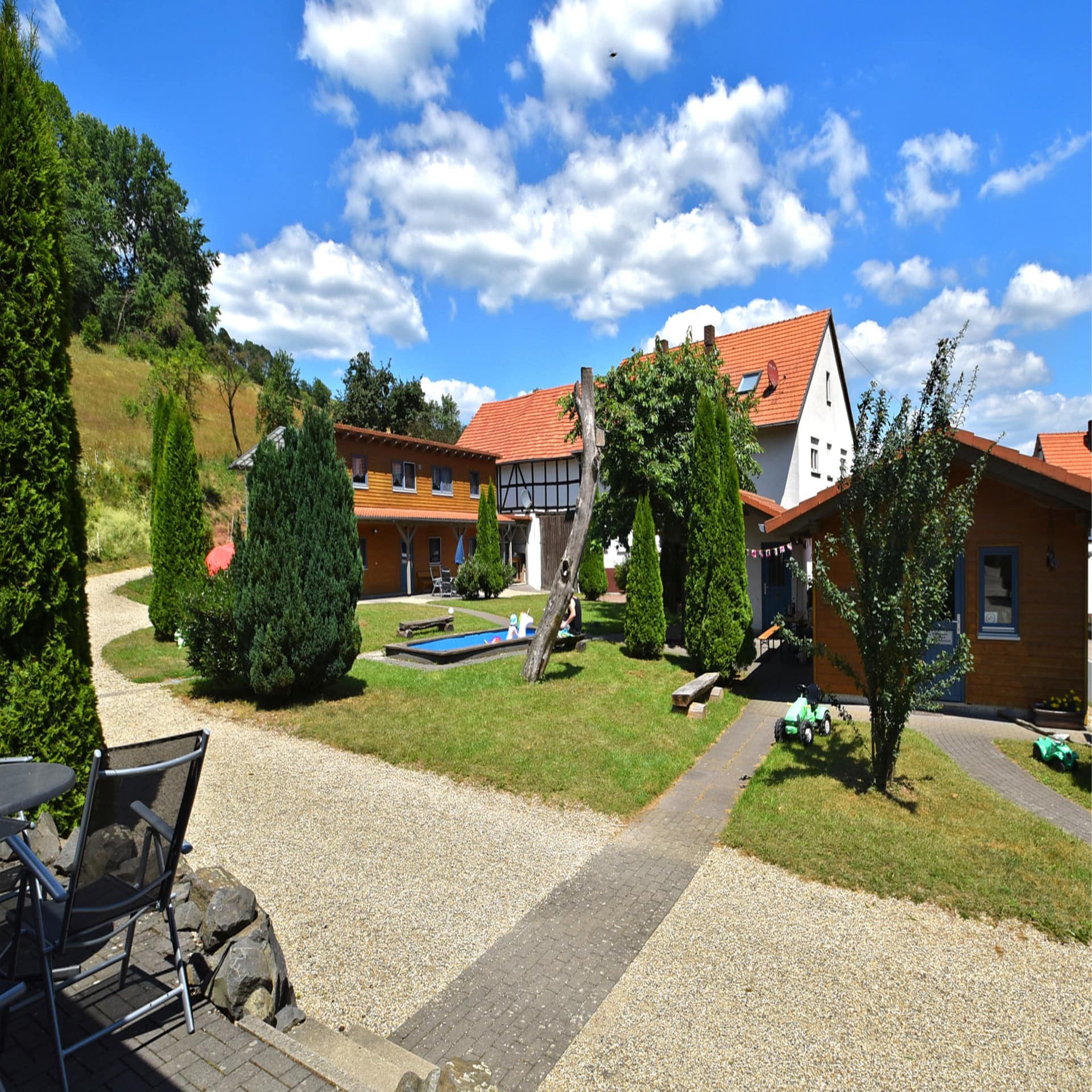 Bauernhof mit Garten in dem sich ein Pool befindet. Die Sonne scheint, der Himmel ist blau. 