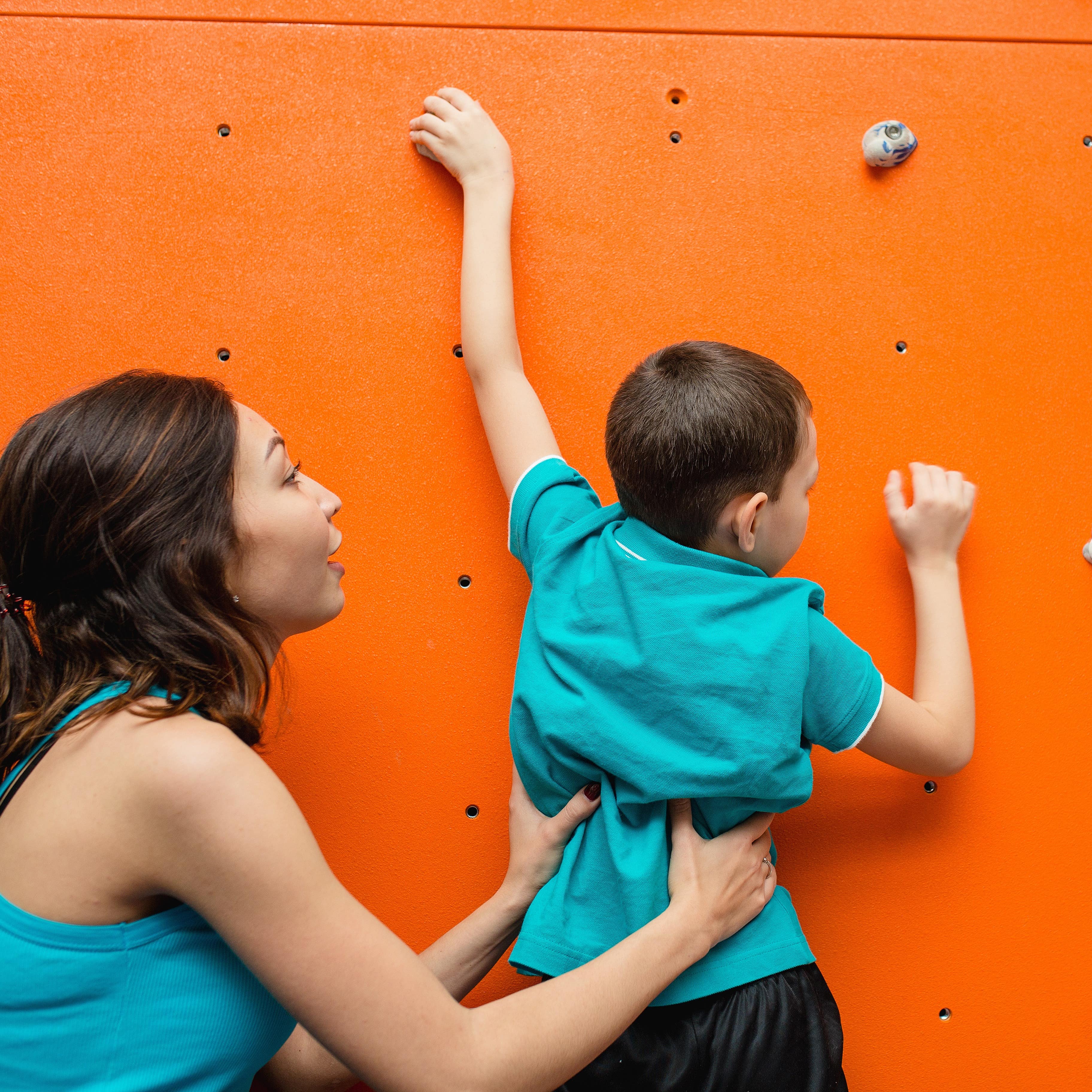 Mutter hilft ihrem Sohn an einer Kletterwand hochzuklettern. Beide tragen türkise Oberteile.