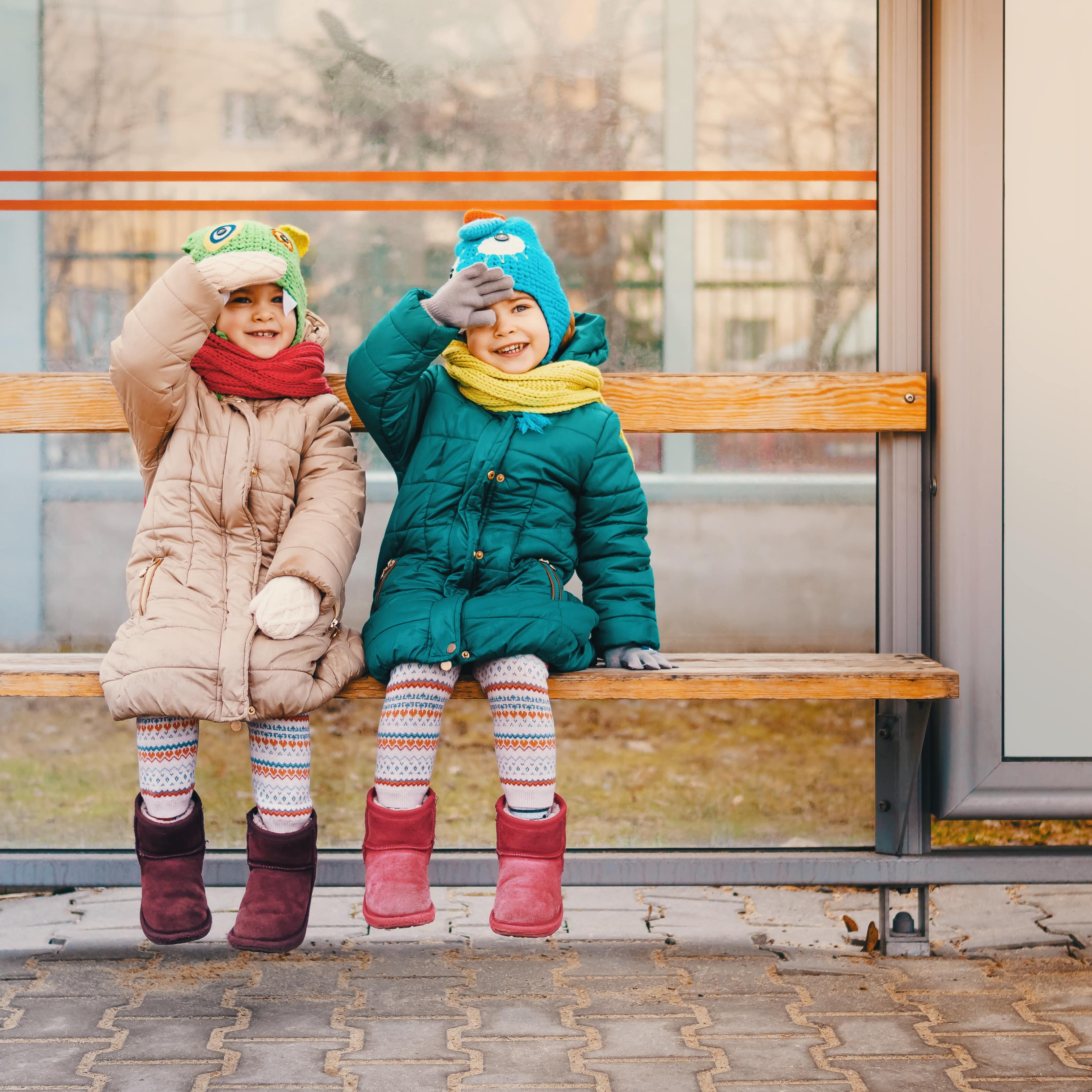 2 kleine Mädchen in Winterkleidung auf einer Bank an einer Bushaltestelle.