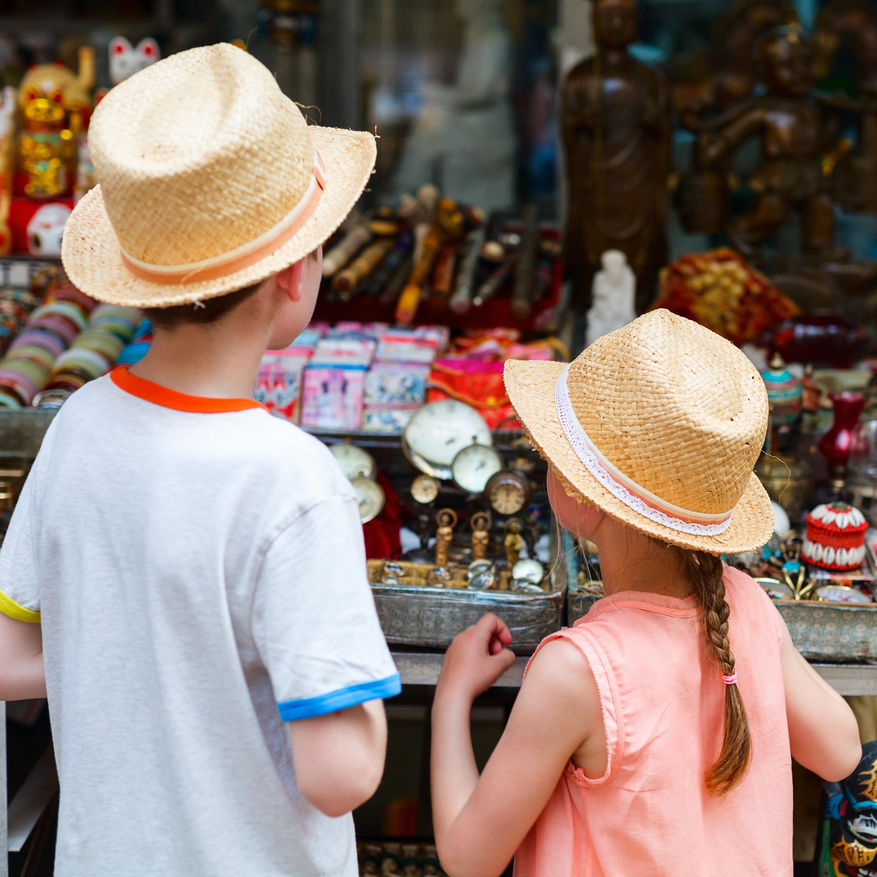 Rückenansicht: 2 Kinder in Sommerkleidung und mit Strohhüten schauen auf die Sachen eines Flohmarktstands. 