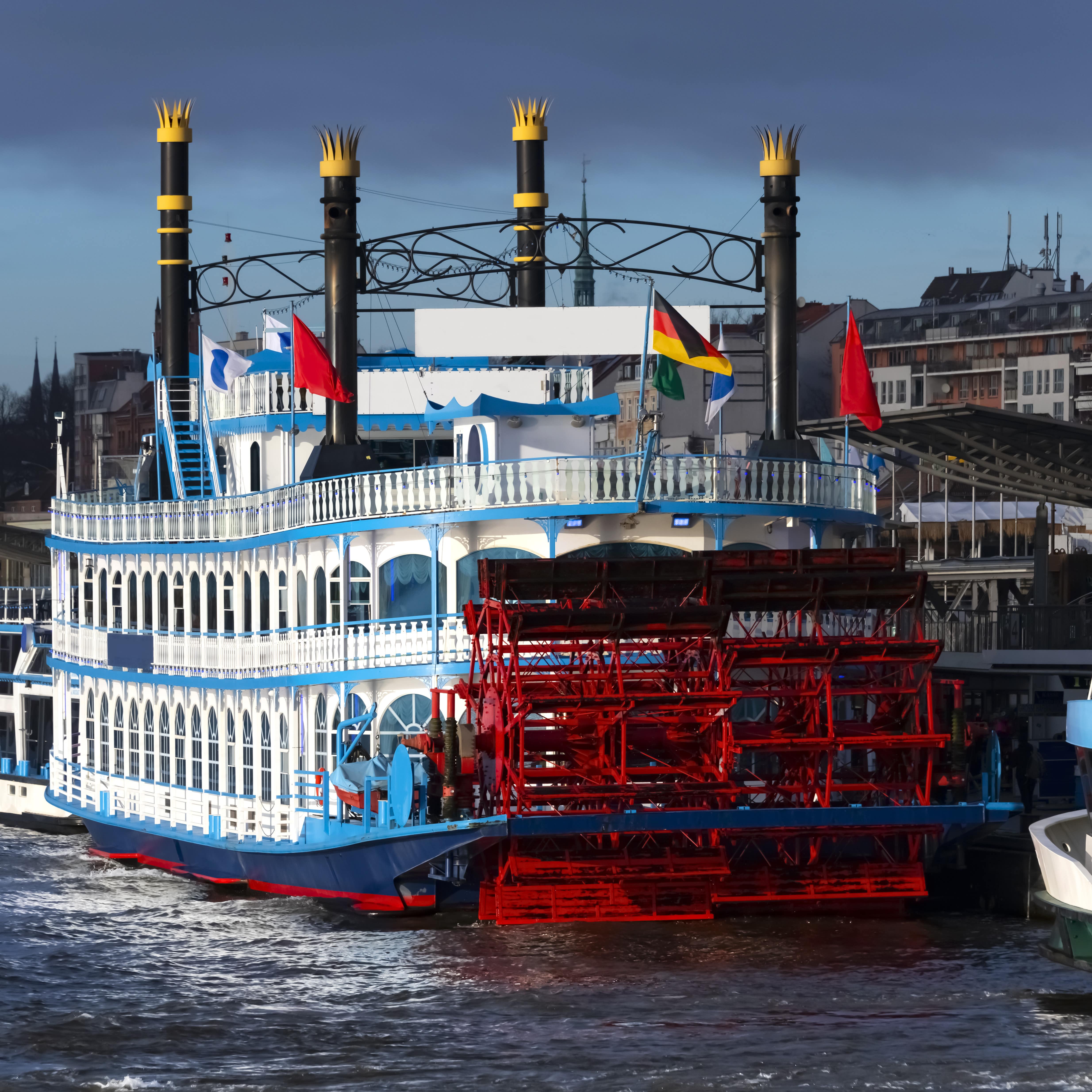 Ein blau-weiß-roter Raddampfer im Hafen von Hamburg. Oben ragen 4 Schornsteine mit gelben Kronen hoch.