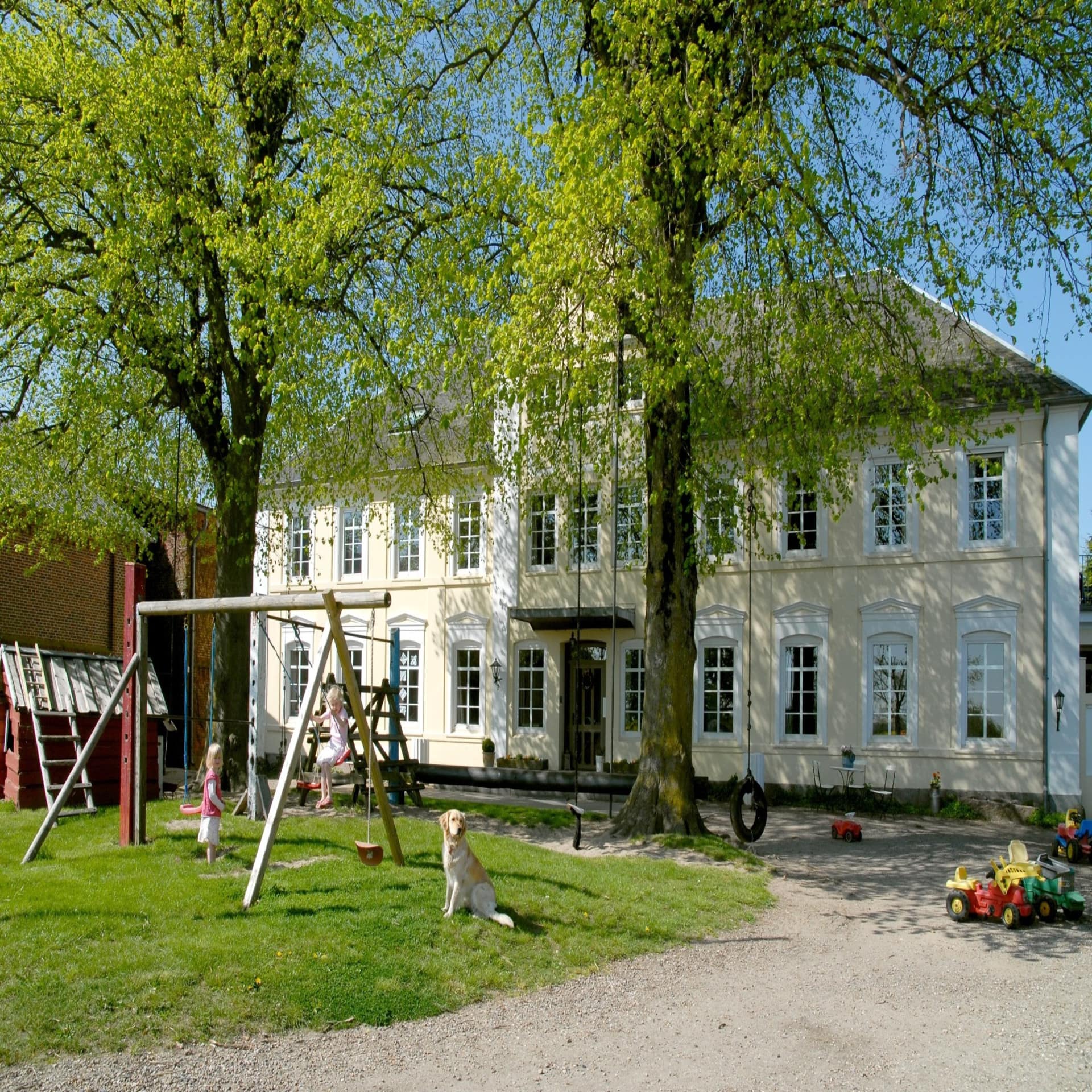 Helles Bauernhaus mit Spielplatz unter Bäumen davor. 2 Kinder, 1 Hund und Spielsachen sind zu sehen. Es ist Frühling.