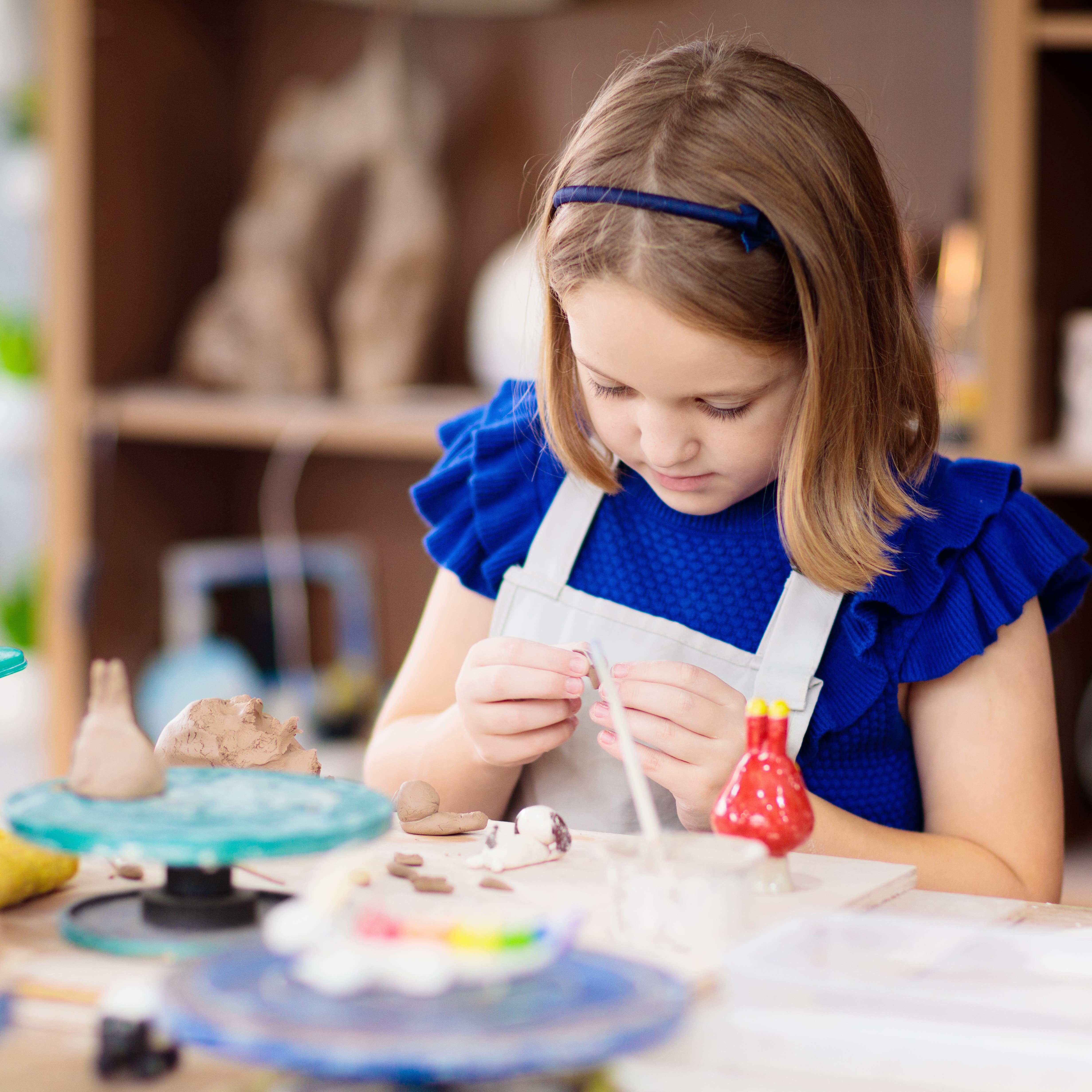 Ein Mädchen in blauem Kleid mit Schürze arbeitet konzentriert beim Töpfern.