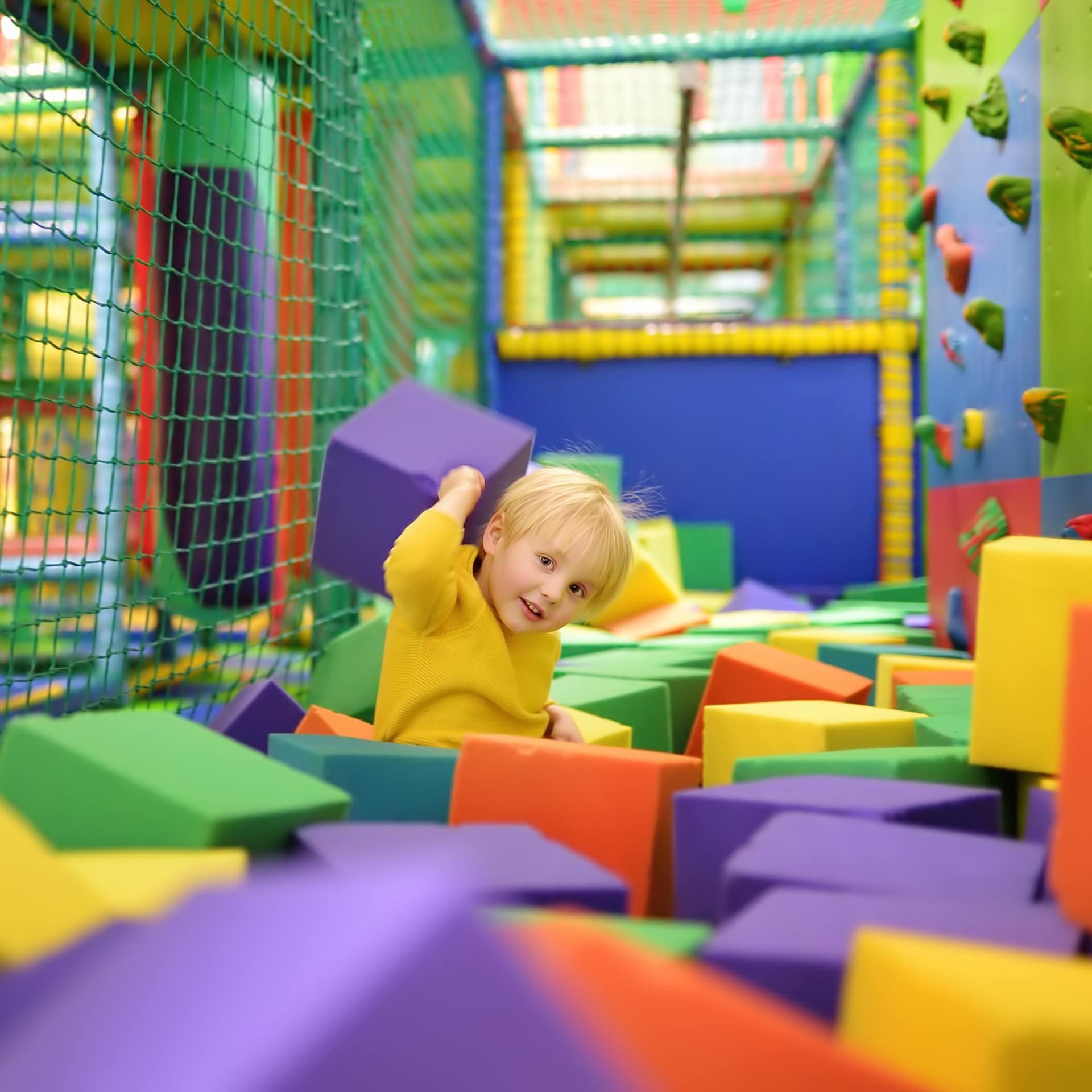 Ein kleiner Junge in gelbem Pulli spielt mit Schaumstoffblöcken in einem Indoor-Spielplatz.