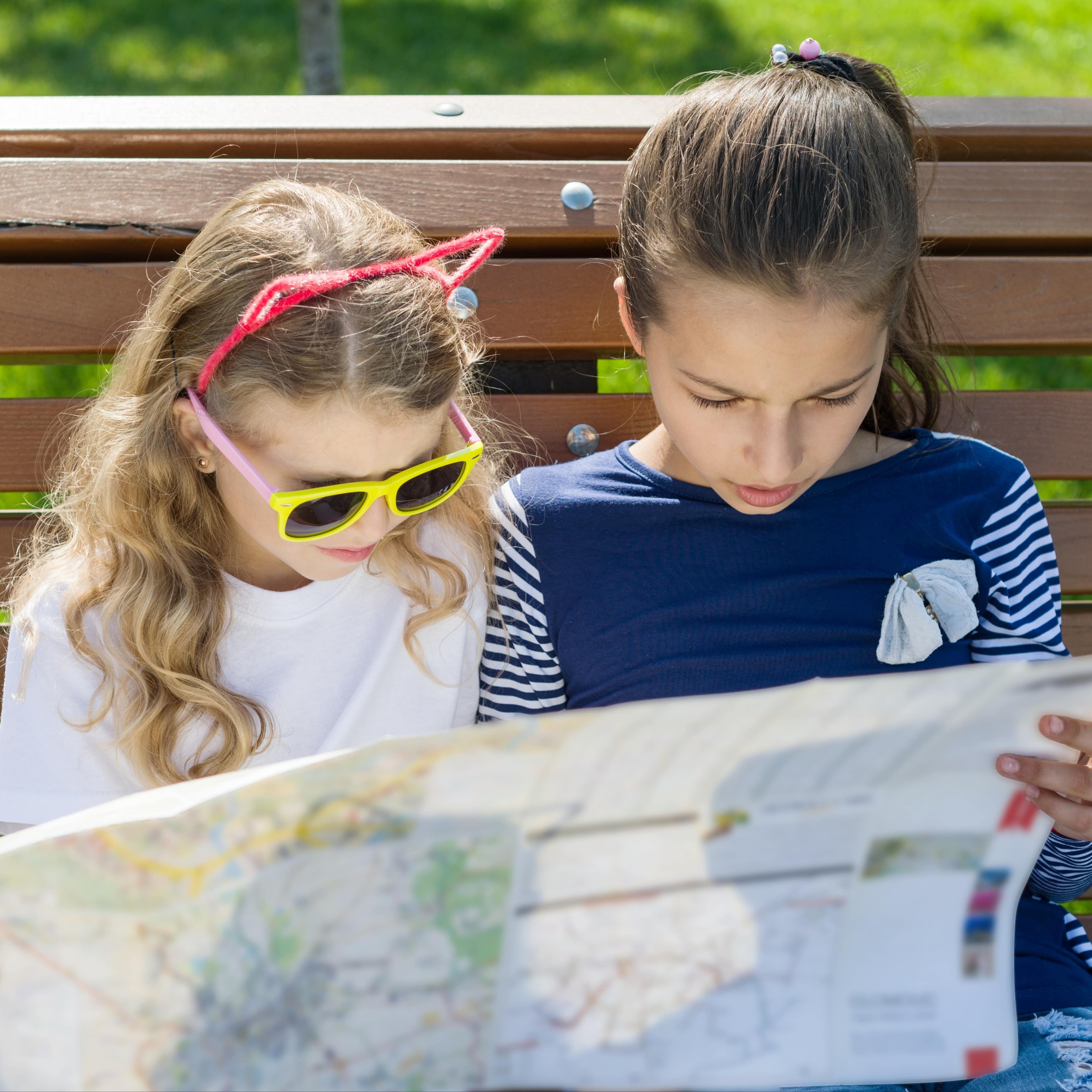 2 Mädchen in Sommerkleidung sitzen auf einer Bank und schauen auf einen Stadtplan.