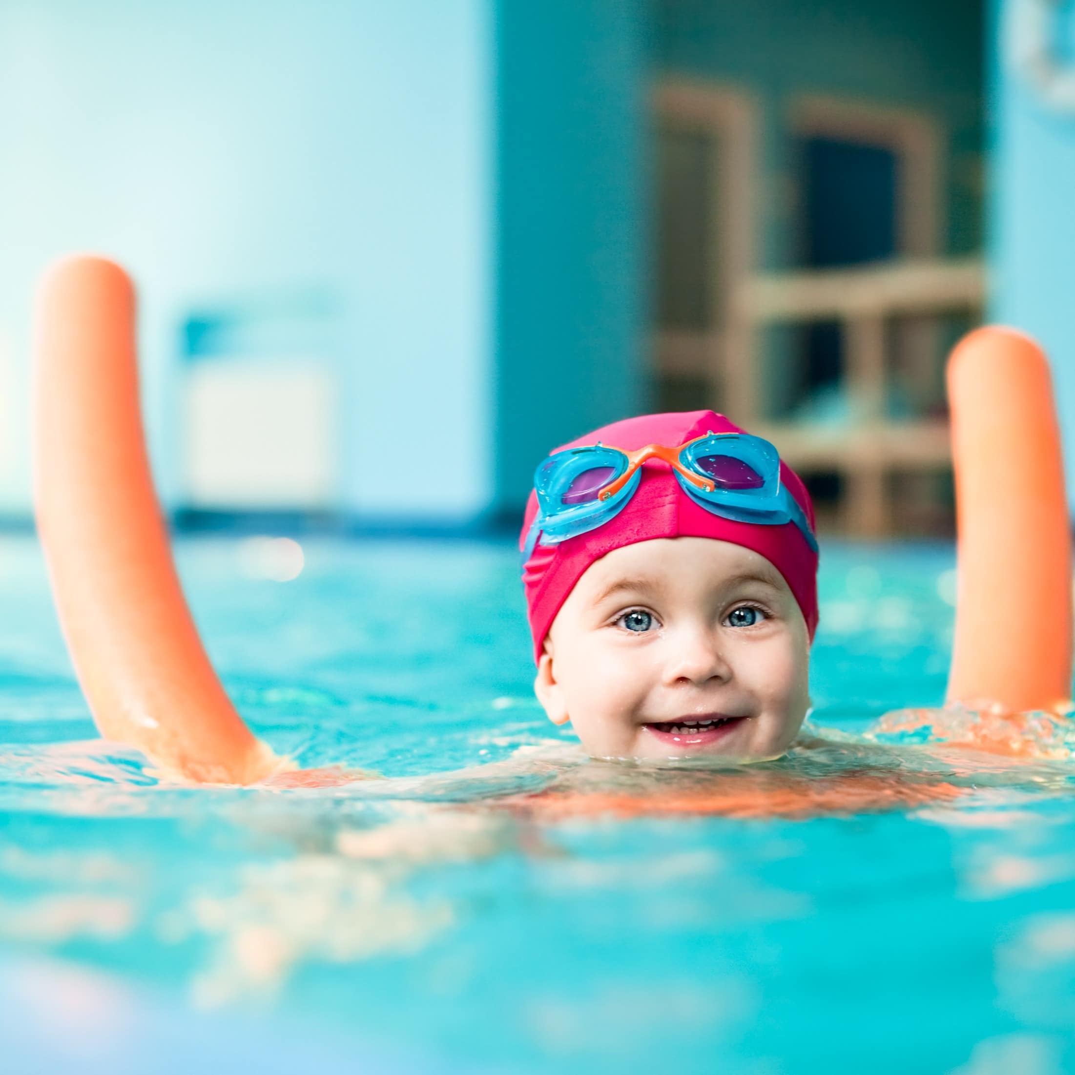 Kleinkind mit pinker Badekappe und Schwimmbrille paddelt mit einer Schwimmnudel durchs Wasser.