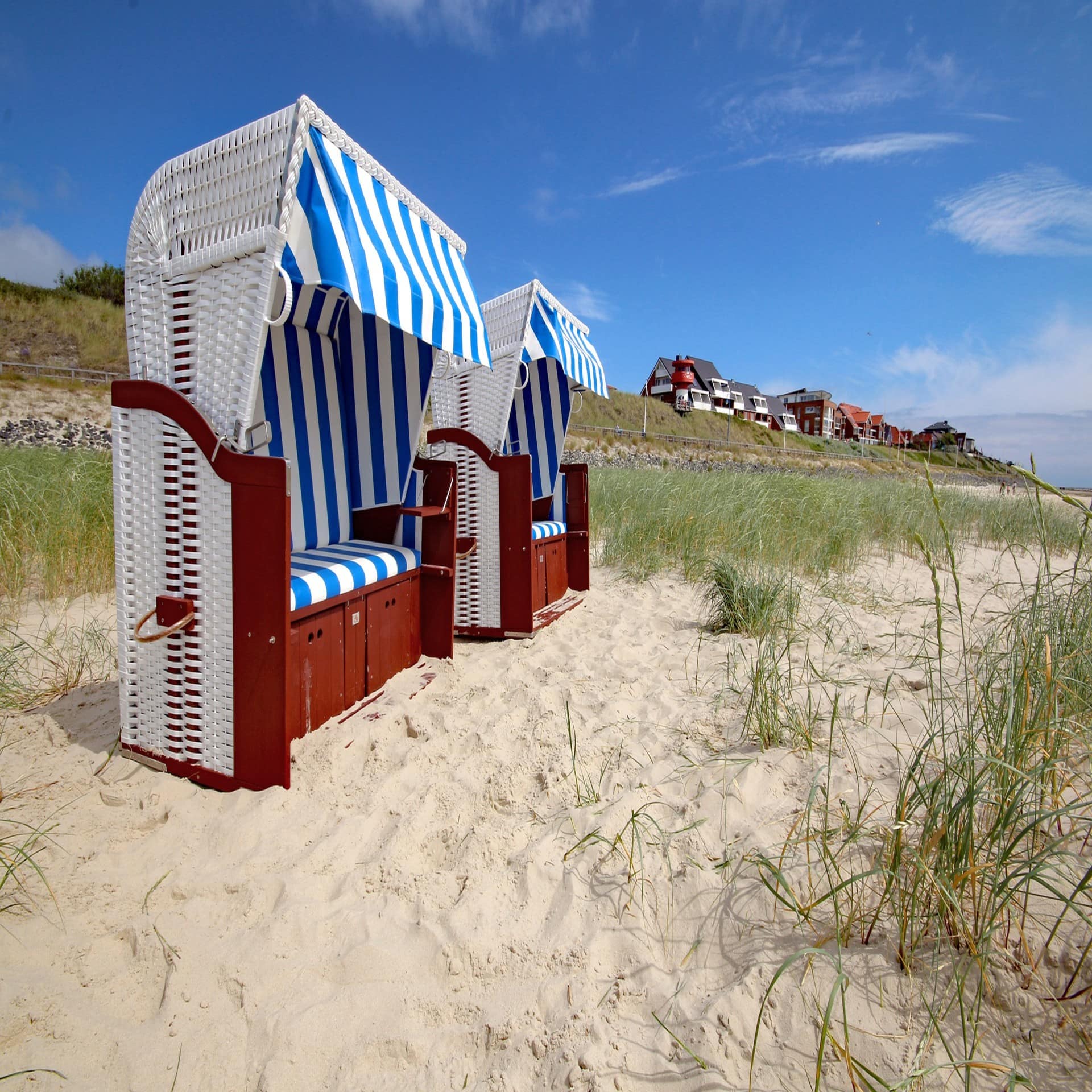 2 blau-weiß gestreifte Strandkörbe im Sand, im Hintergrund die Häuser von Wittdün.
