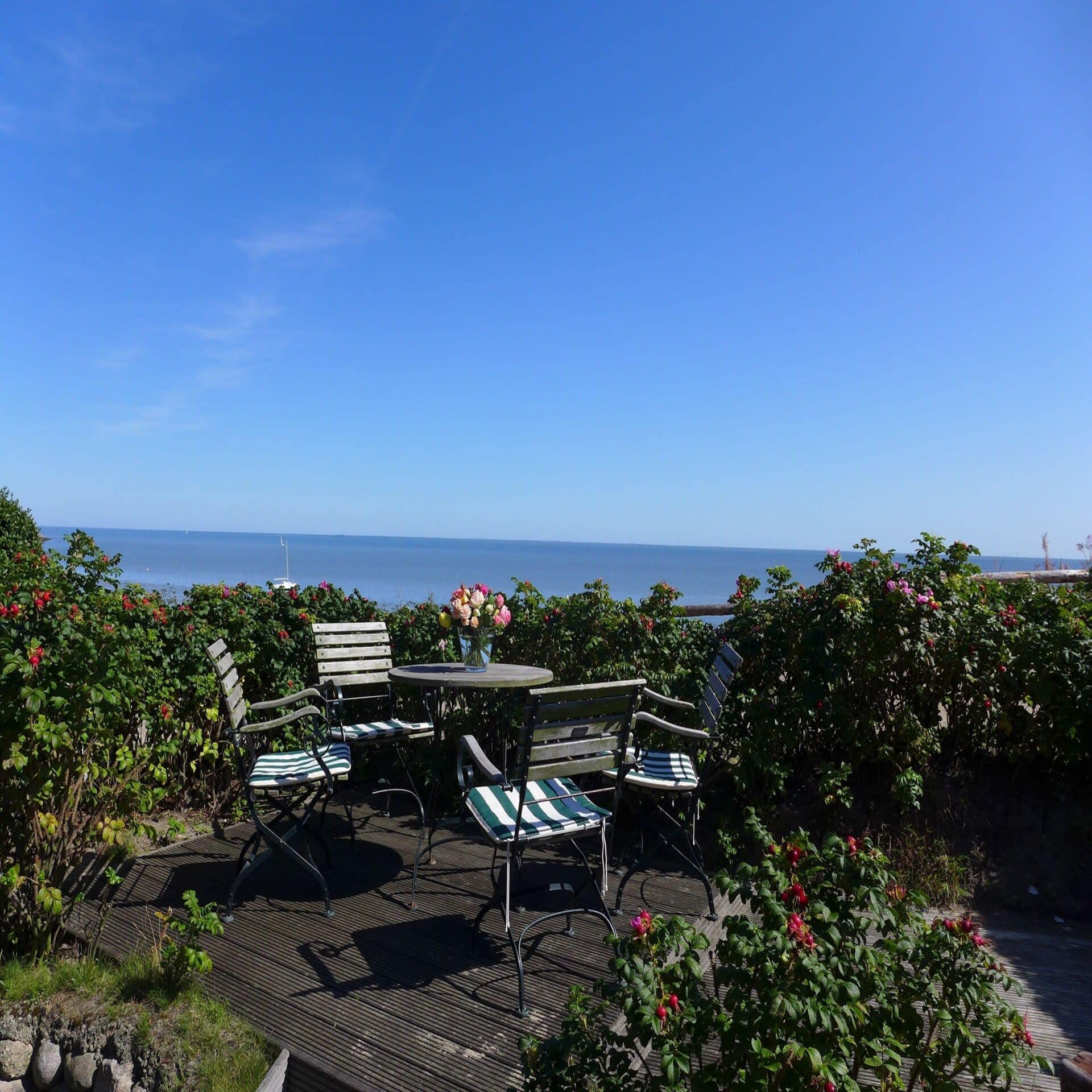 Terrasse mit Tisch und Stühlen in einem Garten mit Blick auf das Meer. 