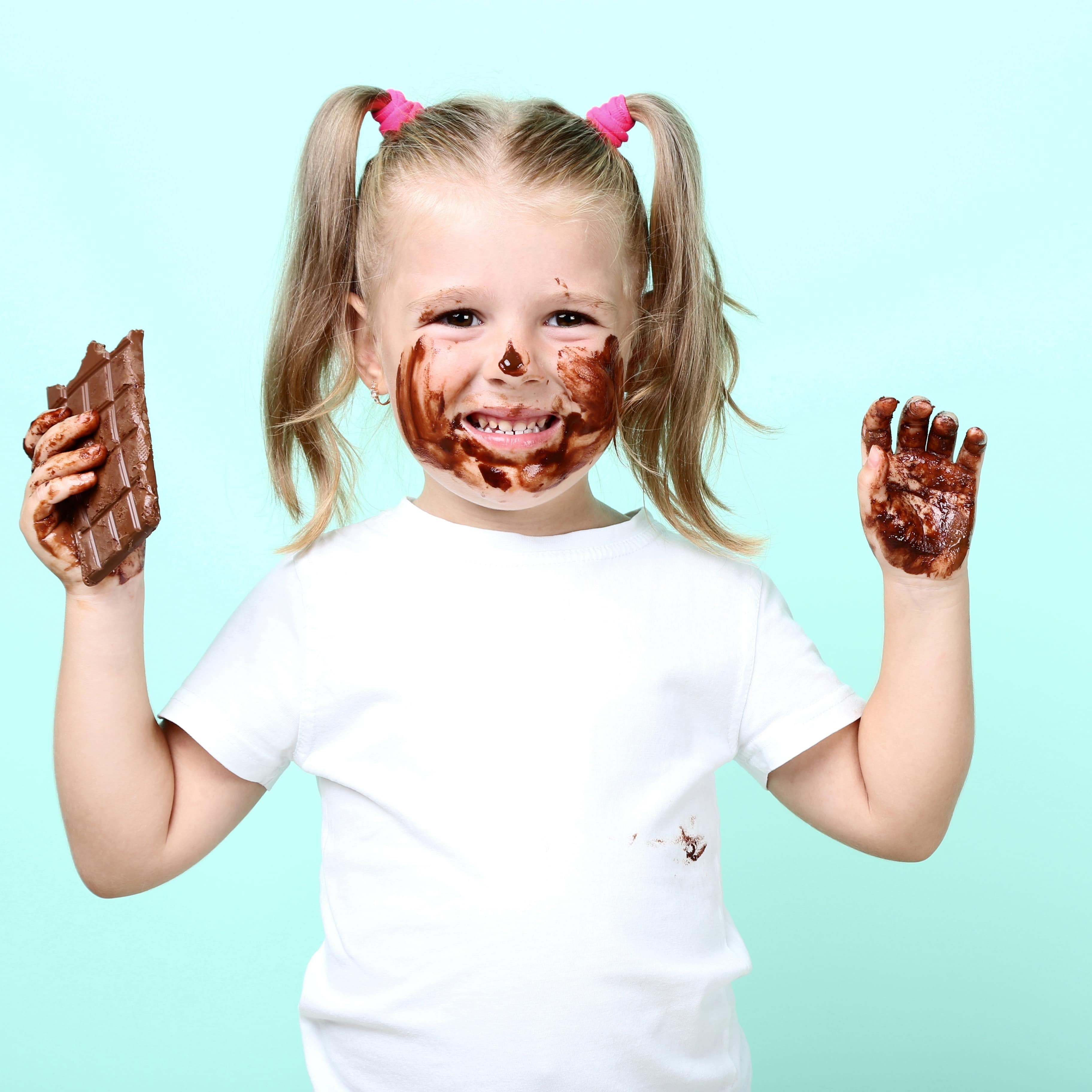 in Mädchen in weißem T-Shirt und einer Tafel Schokolade in der Hand. Sie hat Schokolade auf den Händen und Gesicht. 