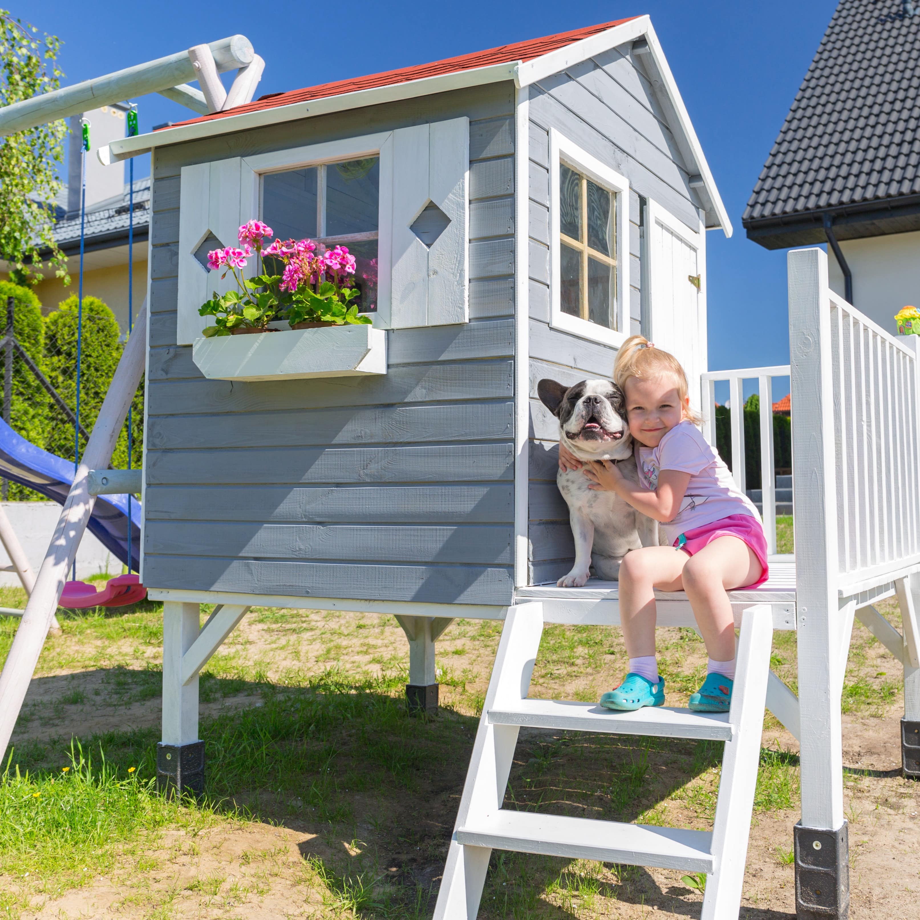 Ein kleines Mädchen und ein Hund sitzen auf der Treppe von einem Spielhaus im Garten.