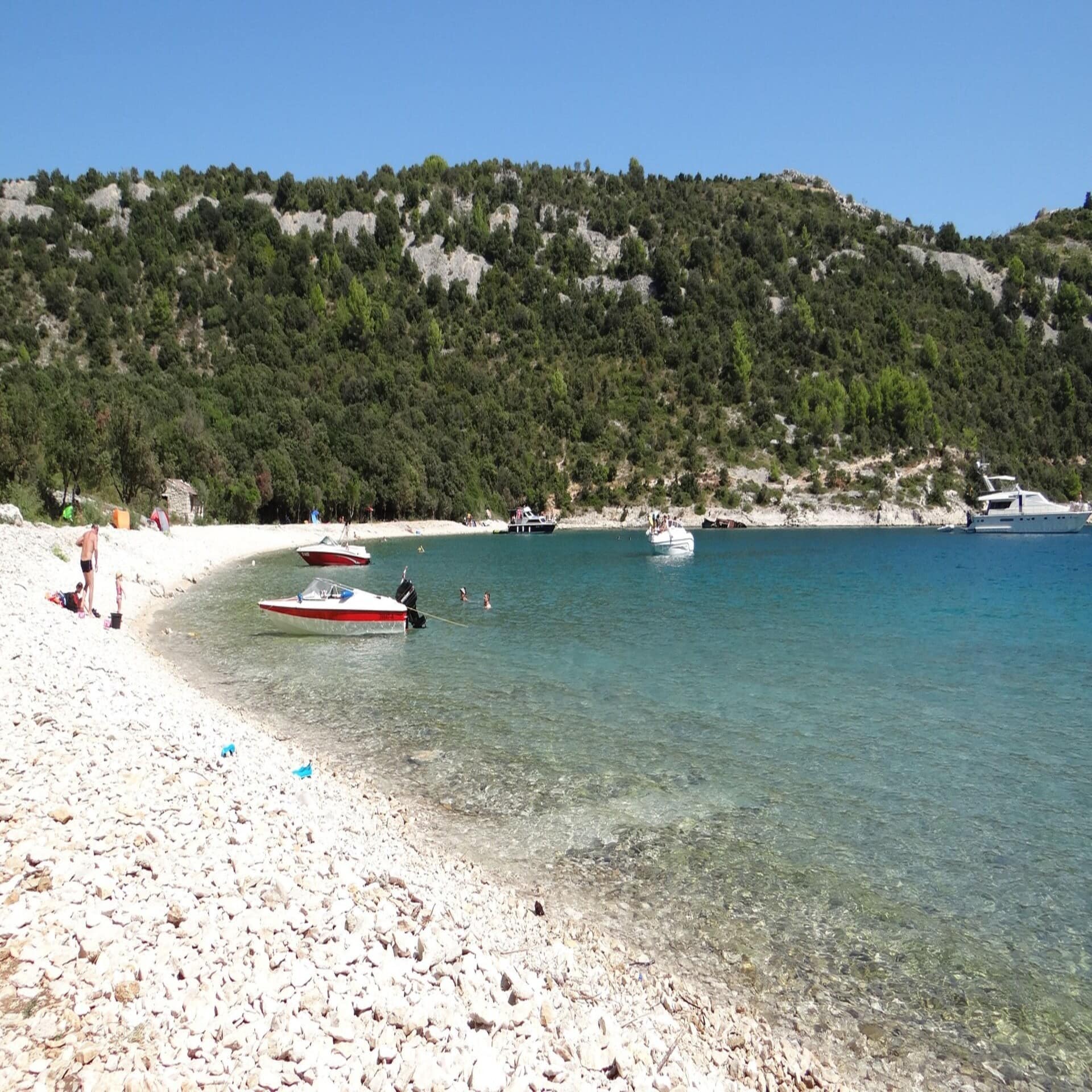 Personen am Kiesstrand, im Wasser einige Boote. Die Sonne scheint.