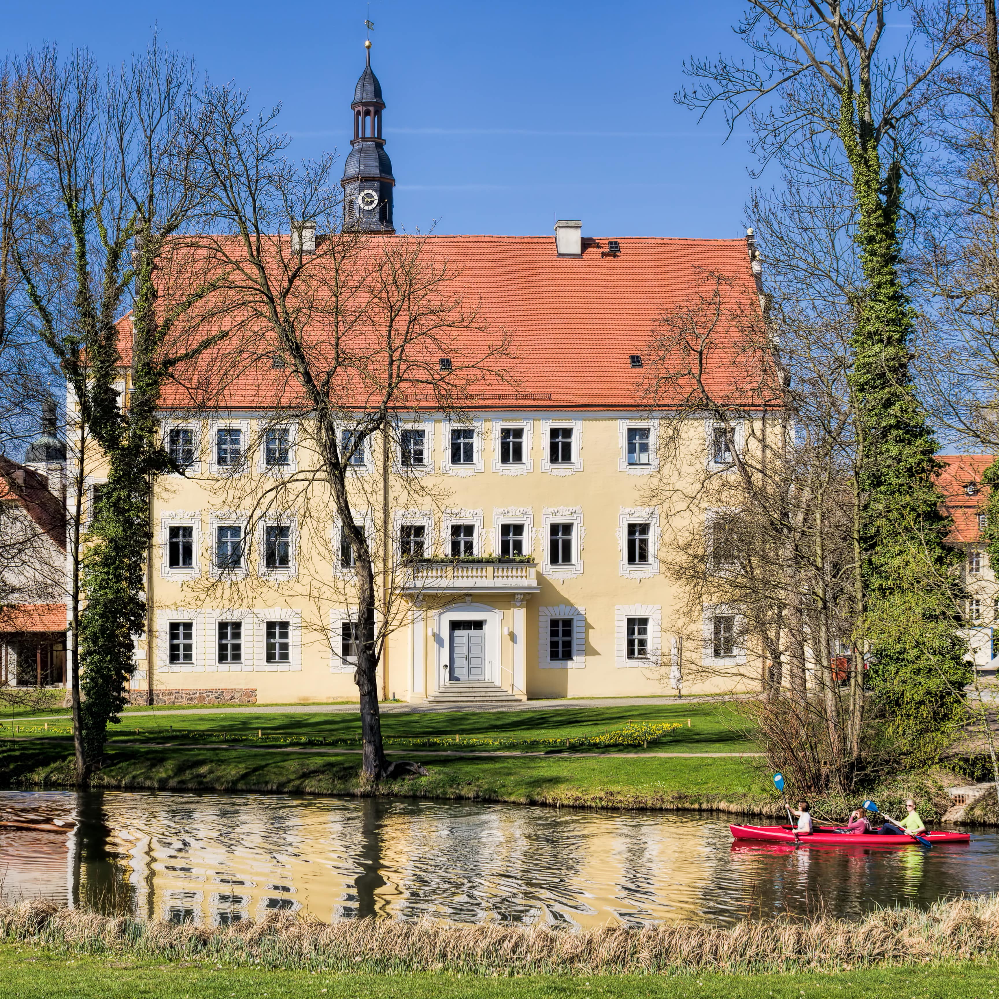 Ein großes sandfarbenes Haus mit rotem Dach, davor eine Wiese und ein Wasserweg auf dem eine Familie im Kanu fährt.