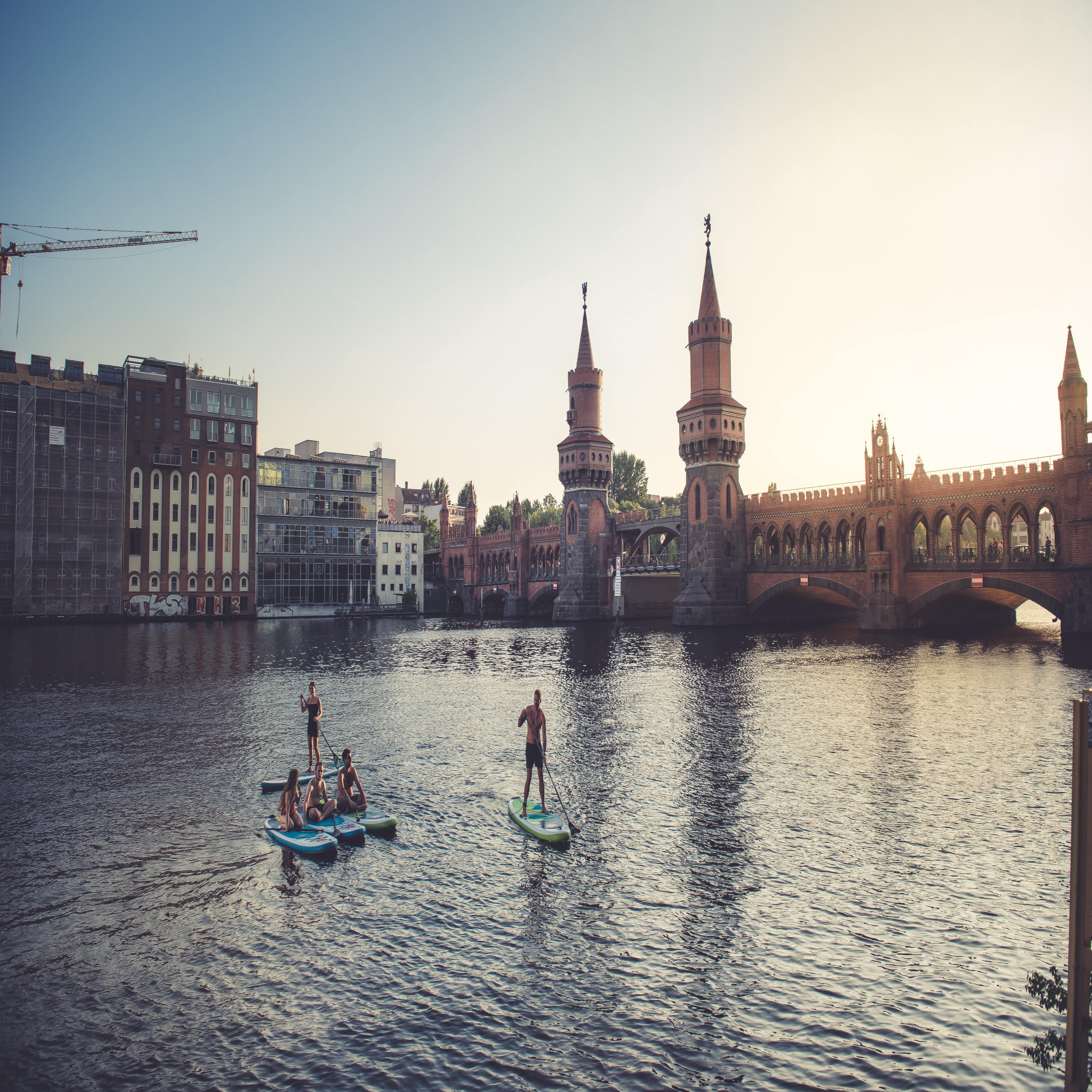 Jugendliche paddeln auf SUPs bei Sonnenuntergang auf der Spree, im Hintergrund eine Brücke und Häuser.