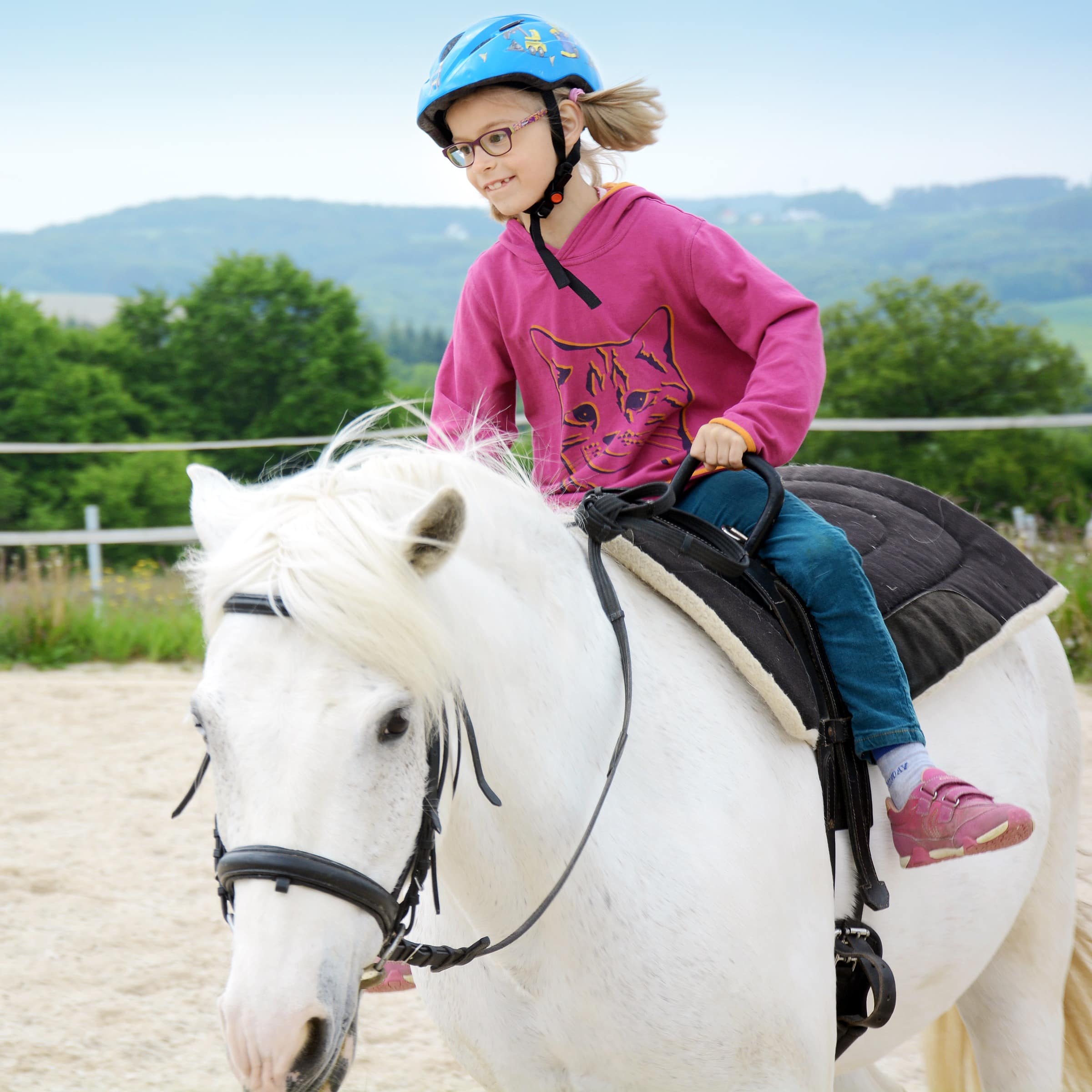 Mädchen mit Brille in pinkem Pulli und Jeans reitet auf einem weißen Pony. 