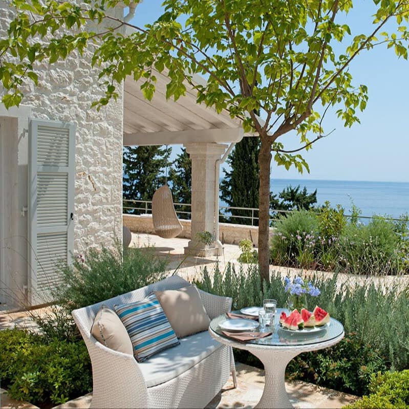 Kleines weißes Ferienhaus auf Korfu mit großer Terrasse, Sitzbank und Tisch mit Melonen darauf sowie Weitblick übers Meer