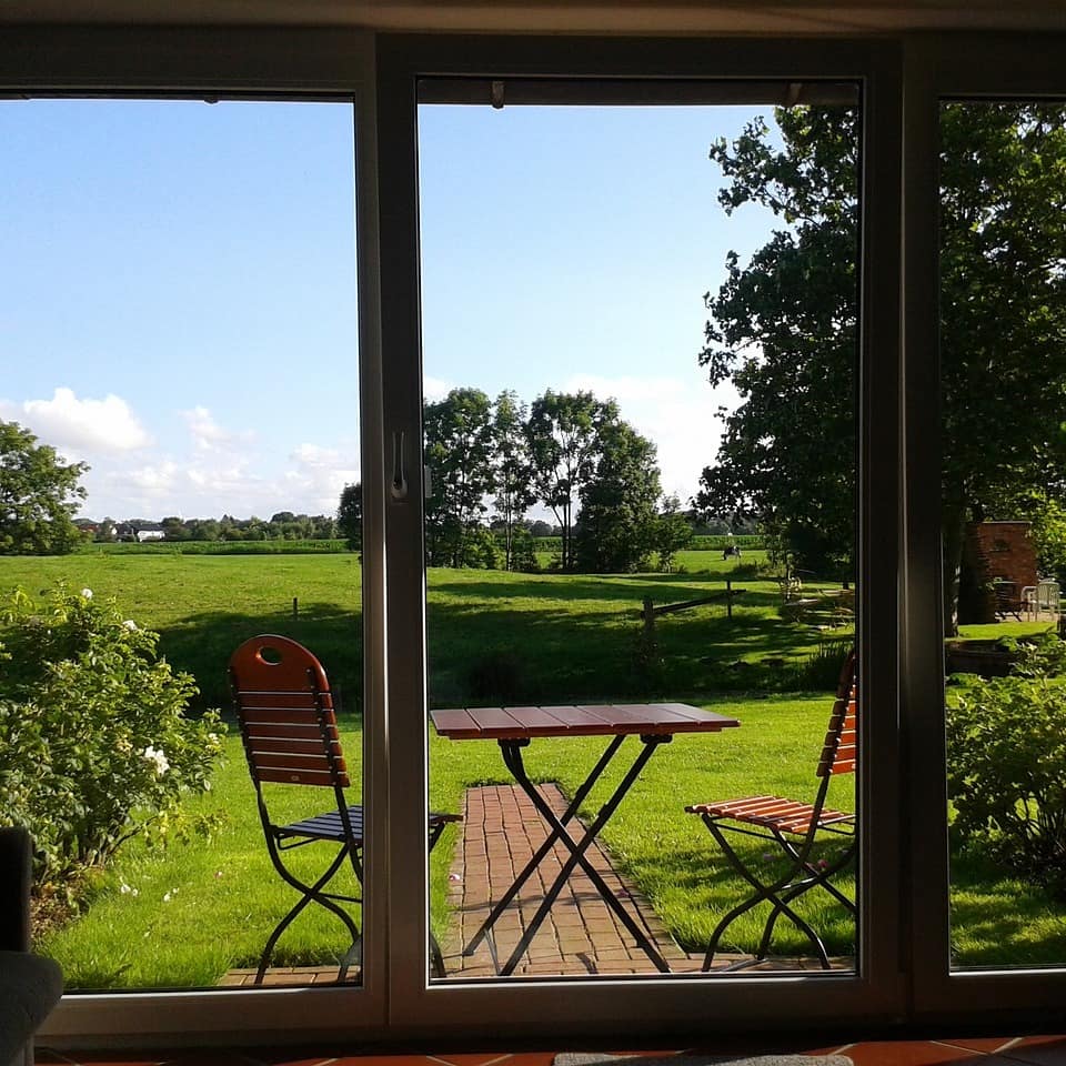 Blick aus dem Fenster eines Hauses auf den Garten mit Tisch und Stühlen sowie das Umland.