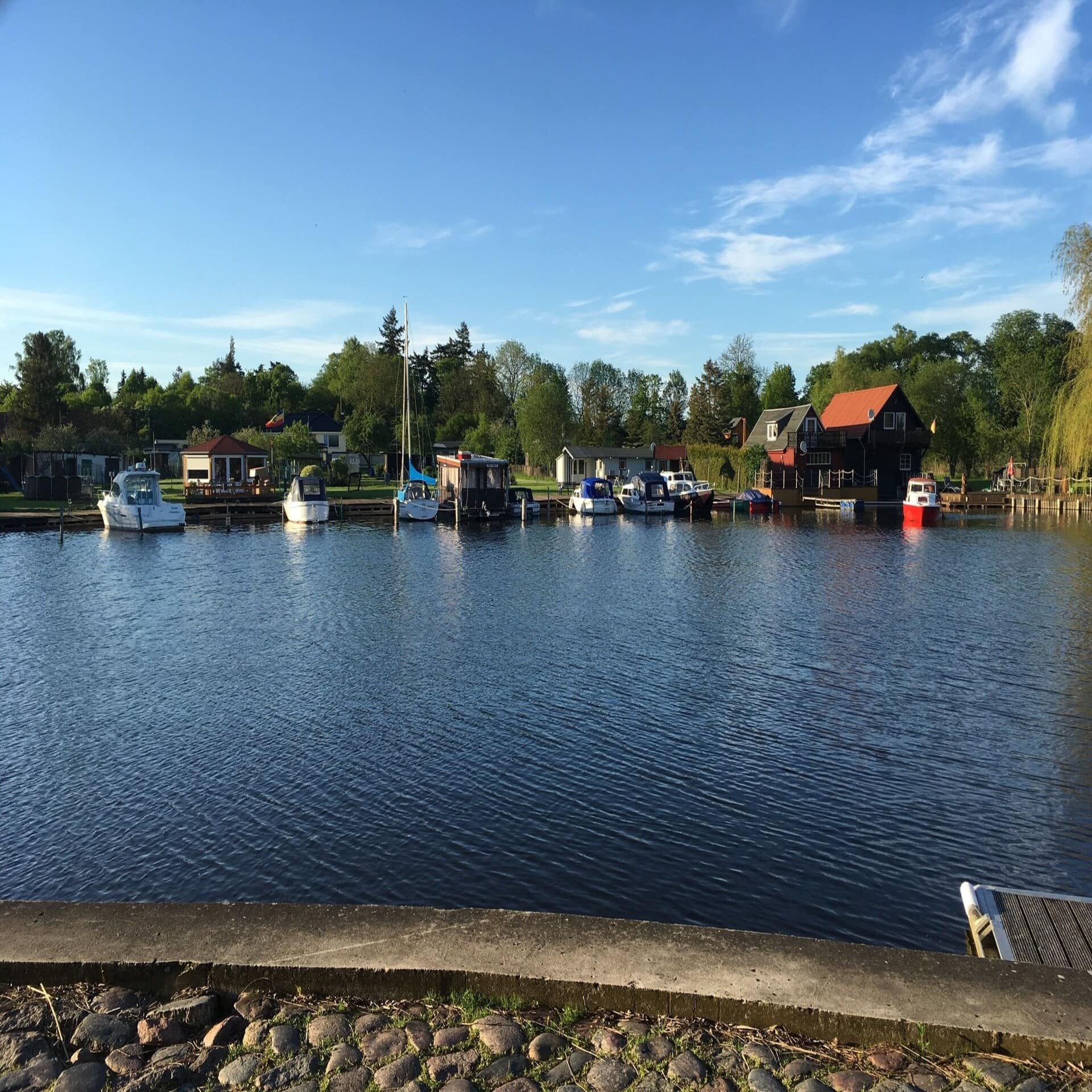 Blick über einen See hinweg auf das gegenüberliegende Ufer: Mehrere Boot vor Anker, dahinter kleine Häuser.