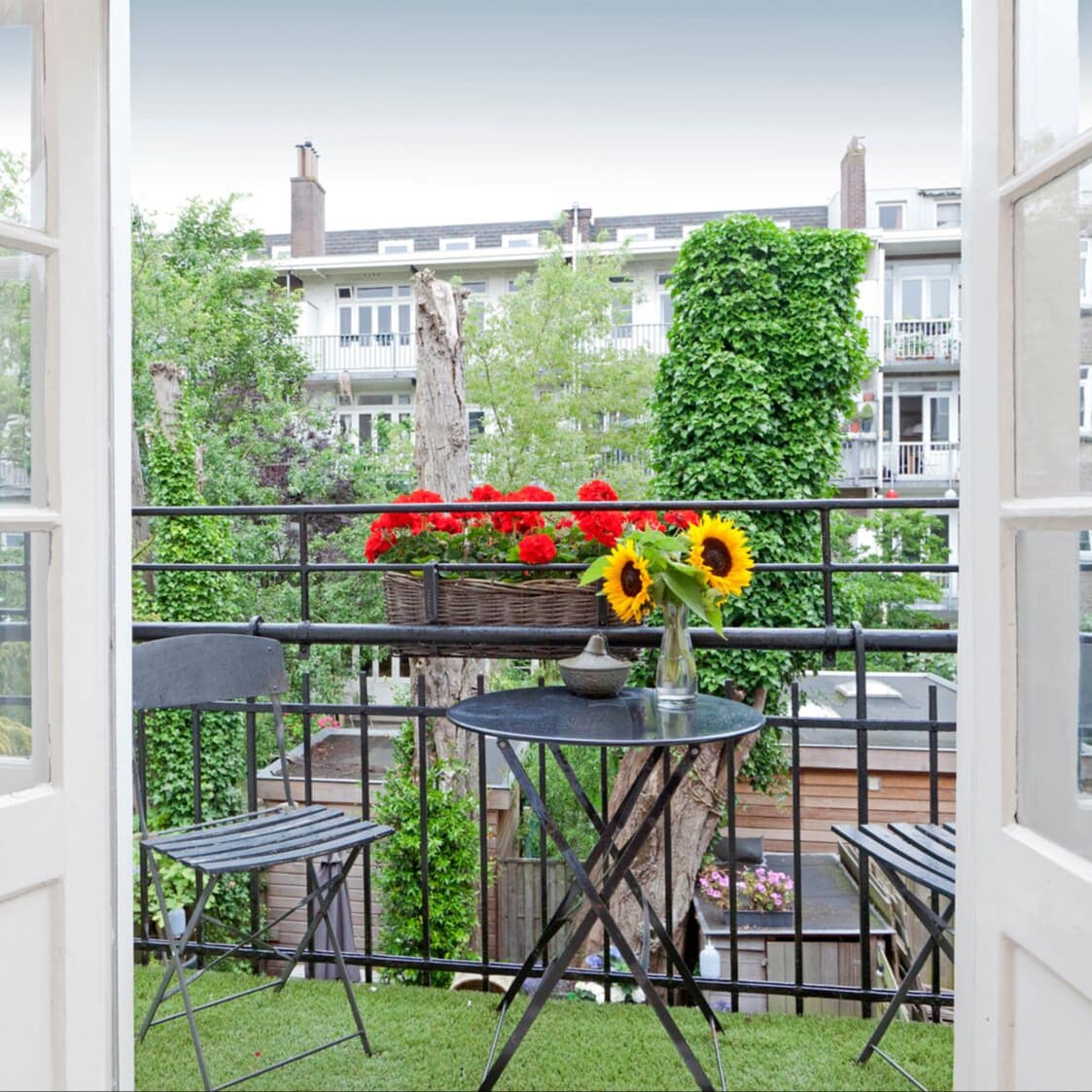 Ferienwohnung in Holland mit hellen Glastüren, die auf einen gemütlichen Balkon mit Tisch, Stühlen und Blumen führen, mit Blick auf Bäume und eine Häuserzeile