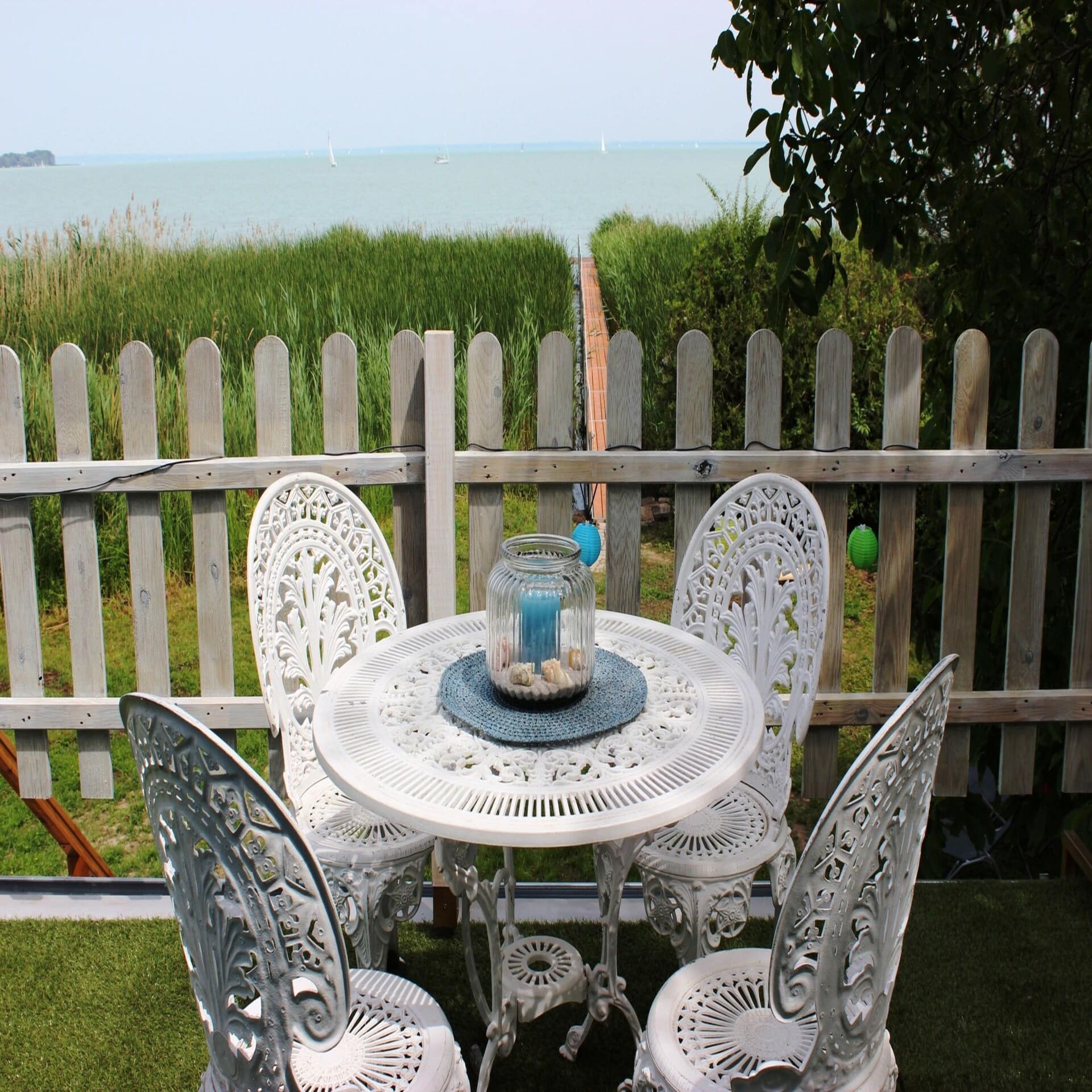 Weißer Tisch und 4 Stühle am Gartenzaun, dahinter ein Schilfgürtel und ein Steg in den Plattensee hinein.