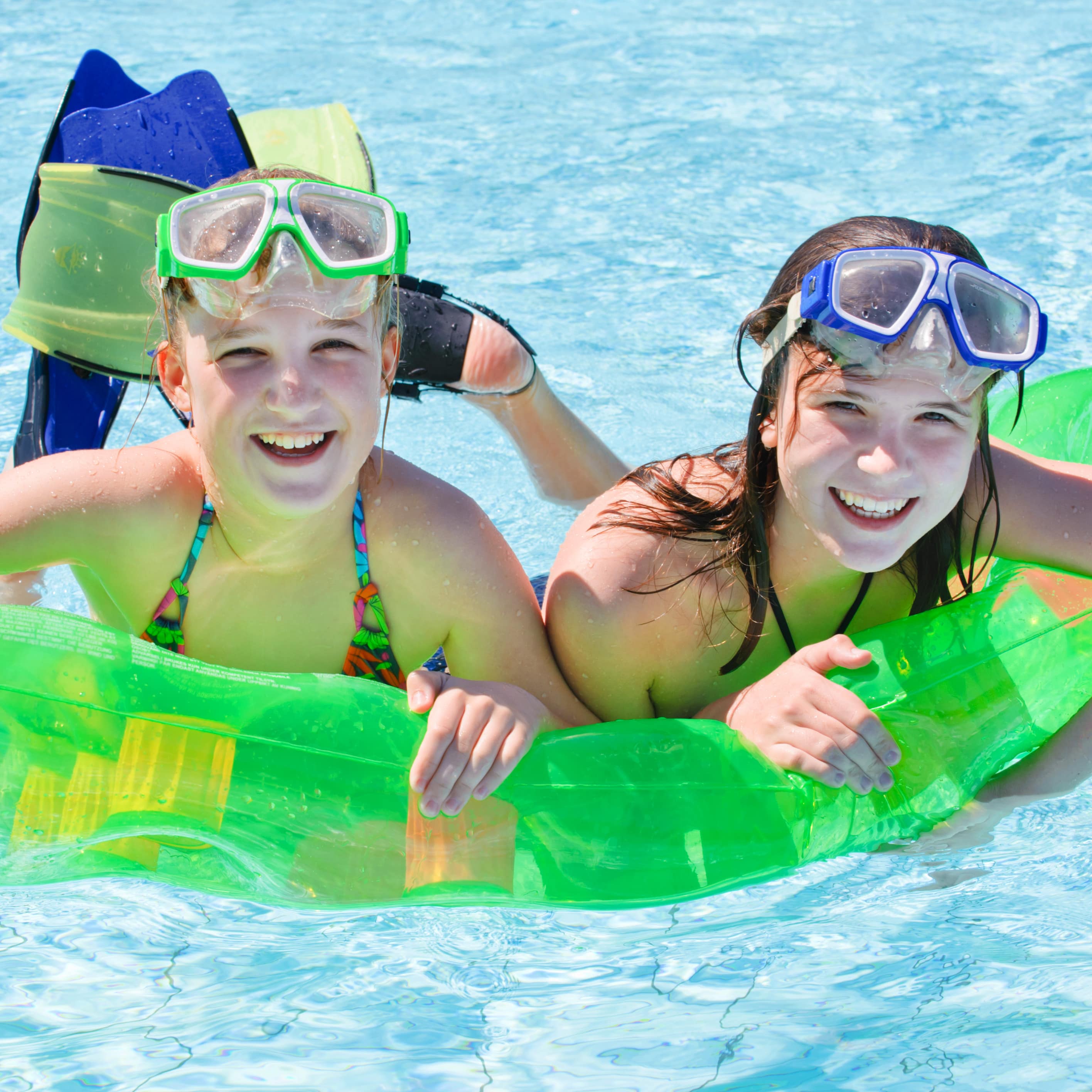 2 Mädchen mit Taucherbrillen auf der Stirn liegen auf einer Luftmatratze im Freibad.