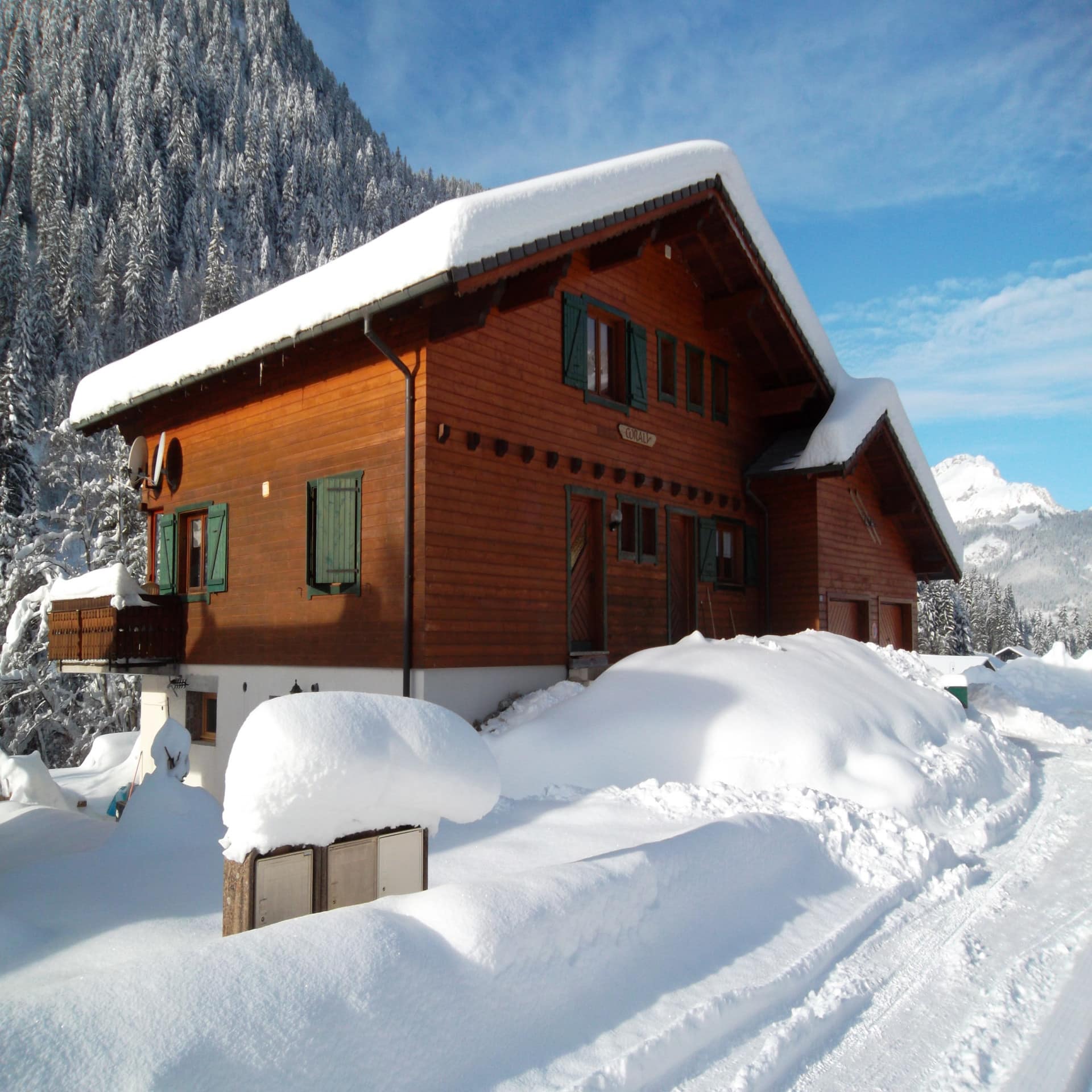 Holzhaus unter einer dicken Schneeschicht in Châtel, die Sonne scheint. Im Hintergrund Bäume.