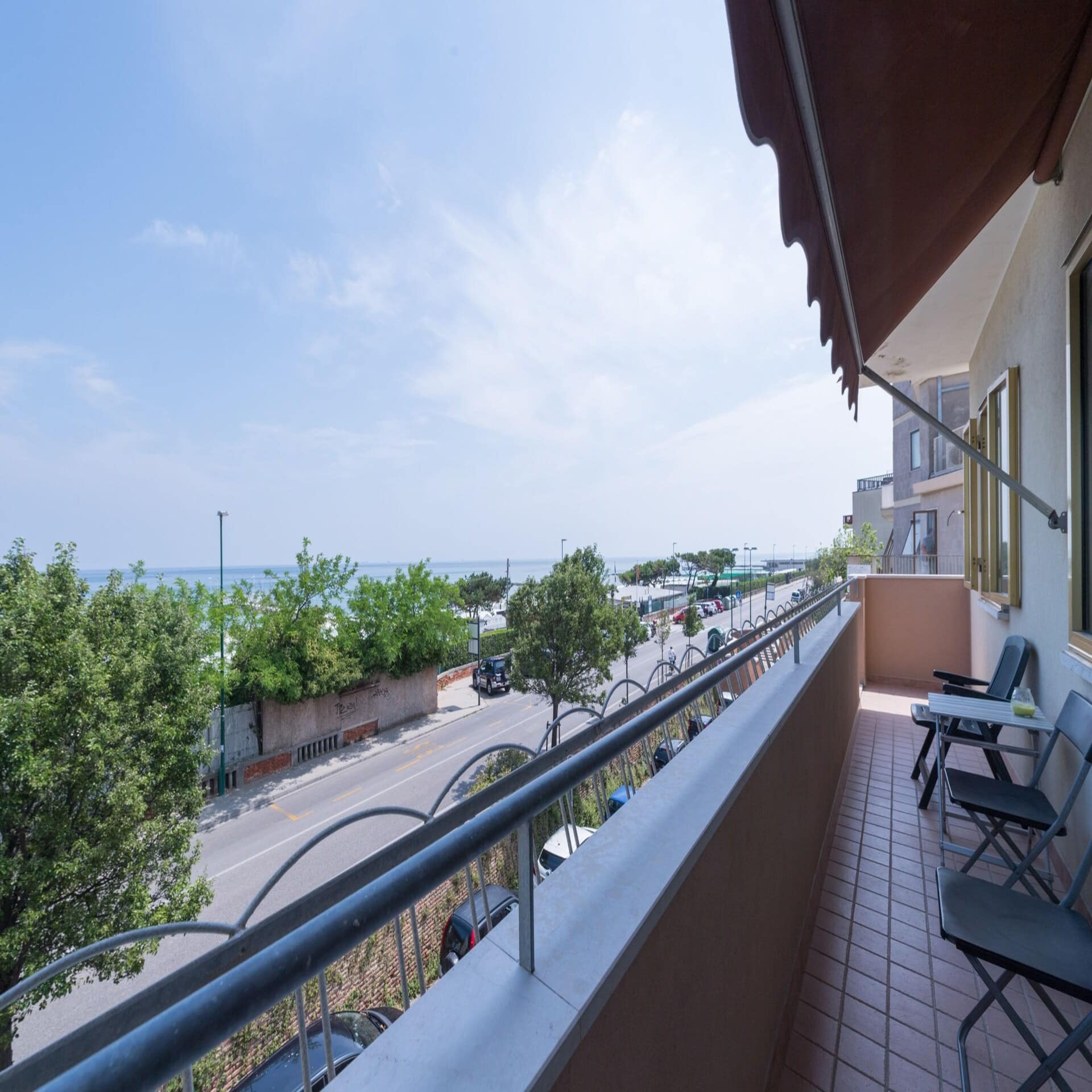 Balkon mit Stühlen mit Blick über eine Straße auf das Meer. Die Sonne scheint.